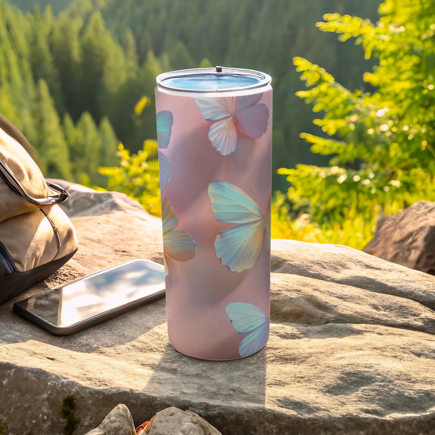 Pink tumbler with pastel butterflies rests on a rock with a phone and backpack.