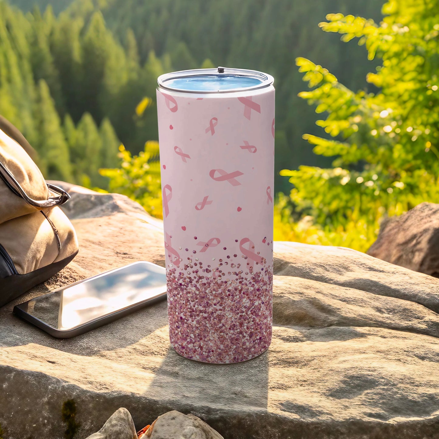 Pink glitter tumbler with pink ribbon pattern, resting on rocks outdoors.