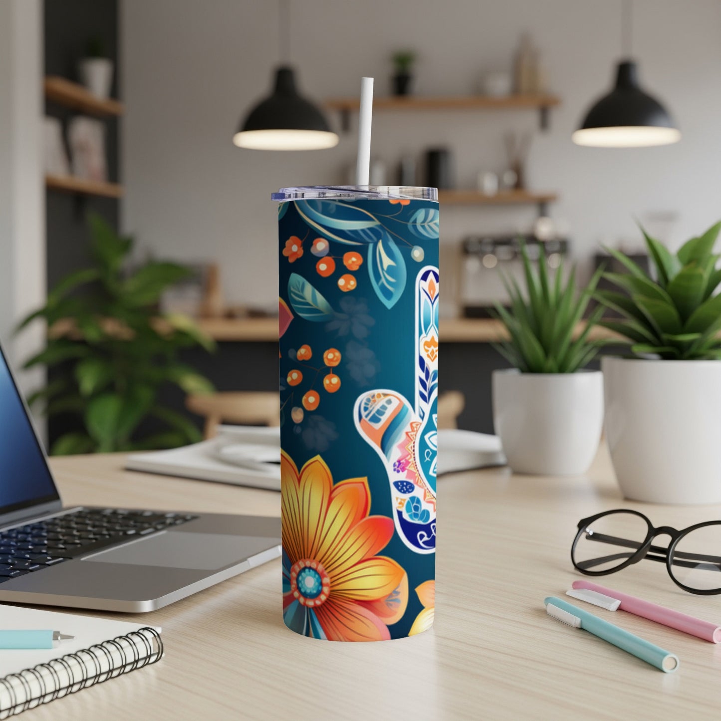 Tall floral tumbler with a straw on a desk with a laptop and pens.