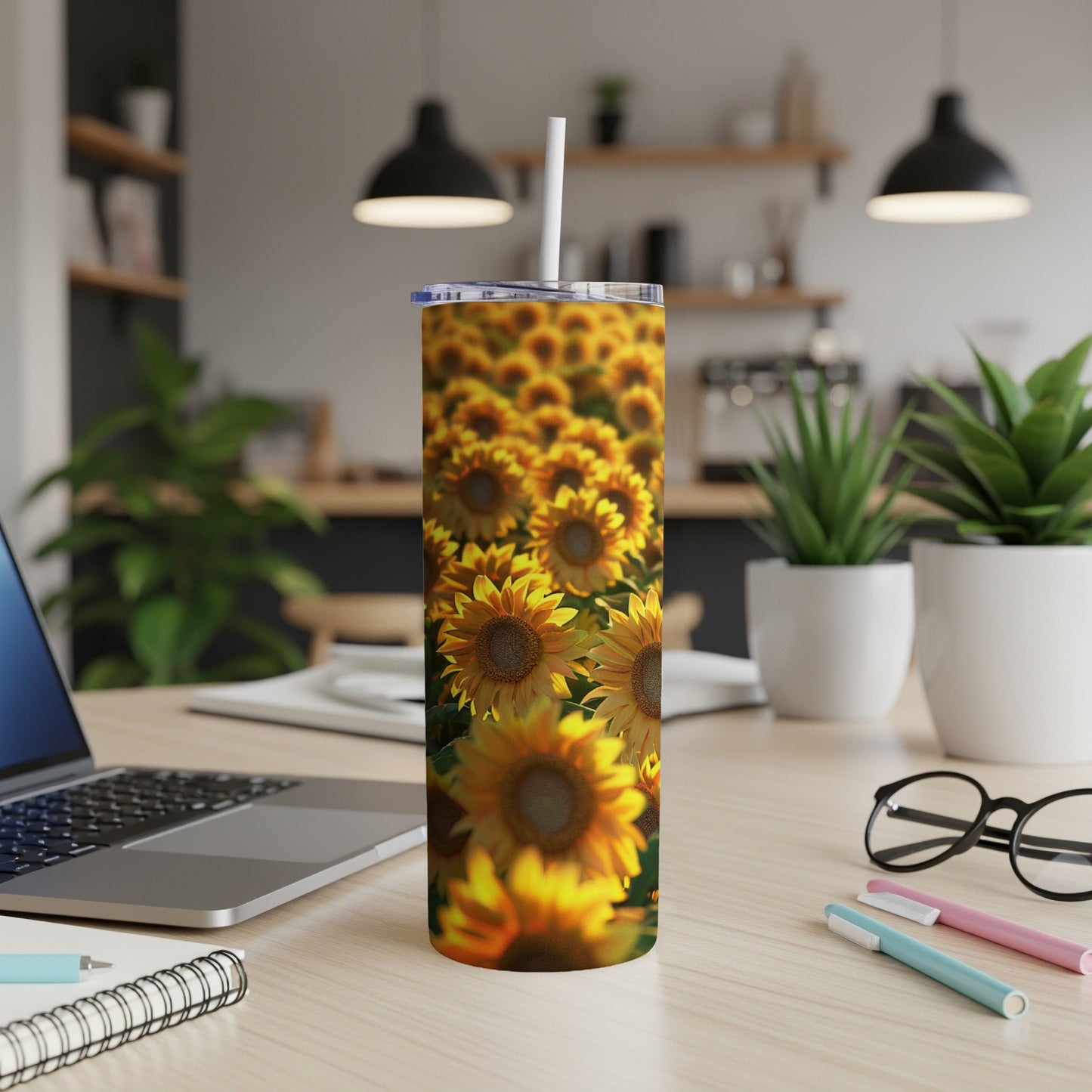 Tall tumbler with sunflowers, white straw, on desk with laptop and pens.