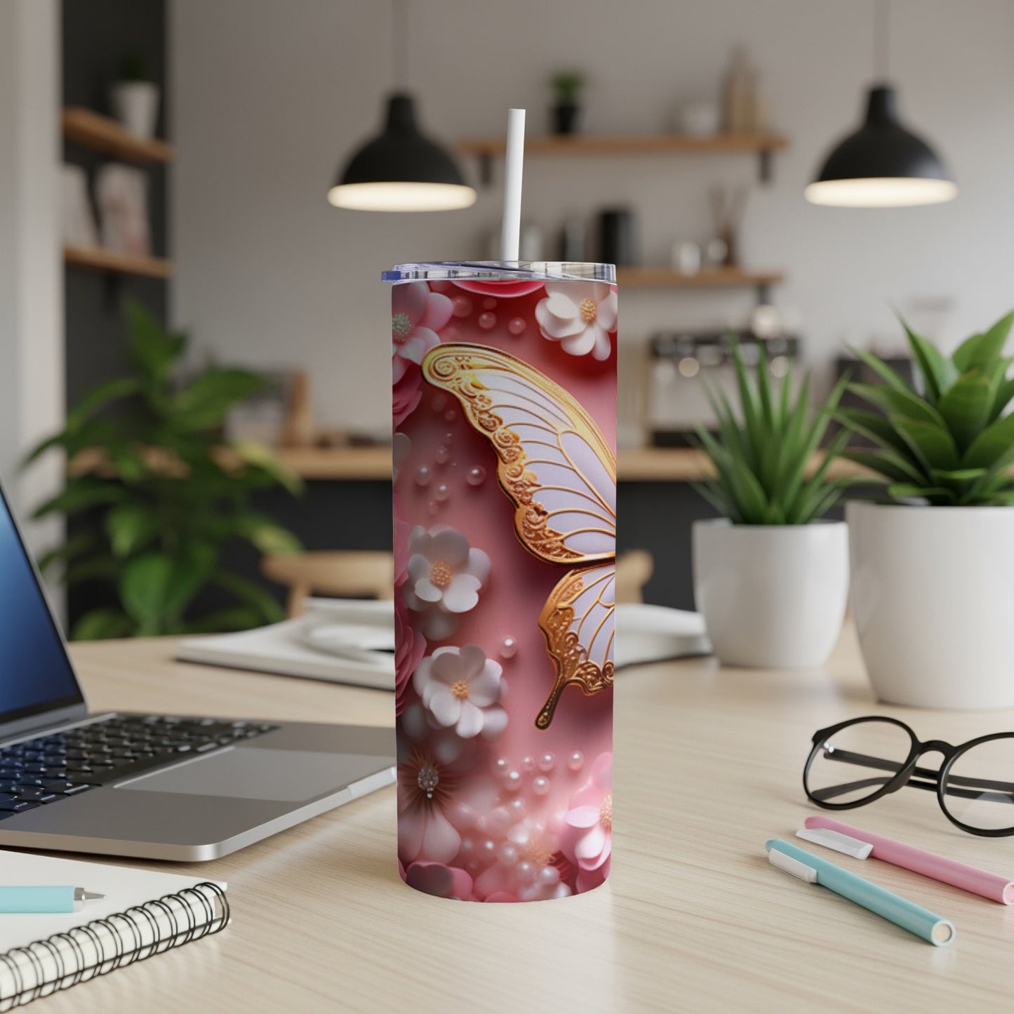 Pink tumbler with butterfly, floral designs, and pearls, with laptop and pens nearby.