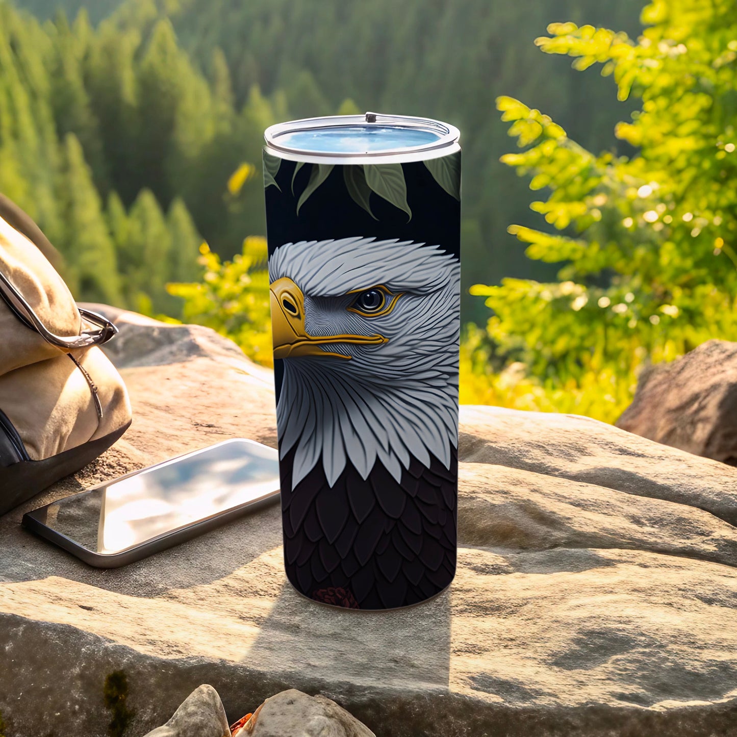 Tall tumbler with eagle design on a rock with phone and bag.