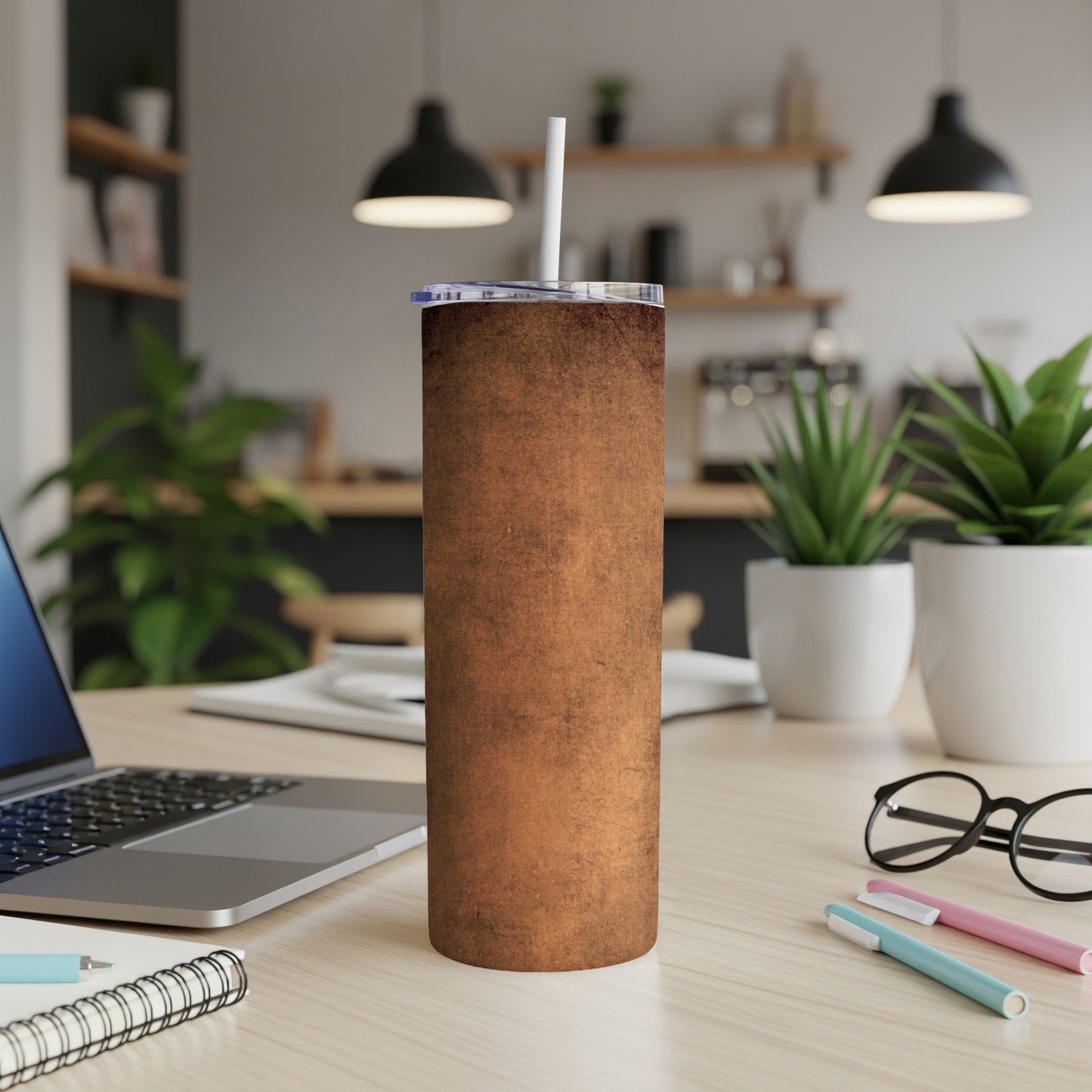 Brown distressed tumbler with straw on wooden desk near laptop and plants.