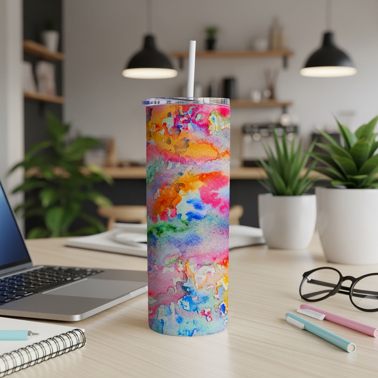 Tall tumbler with colorful watercolor design, on a desk with laptop and pens.