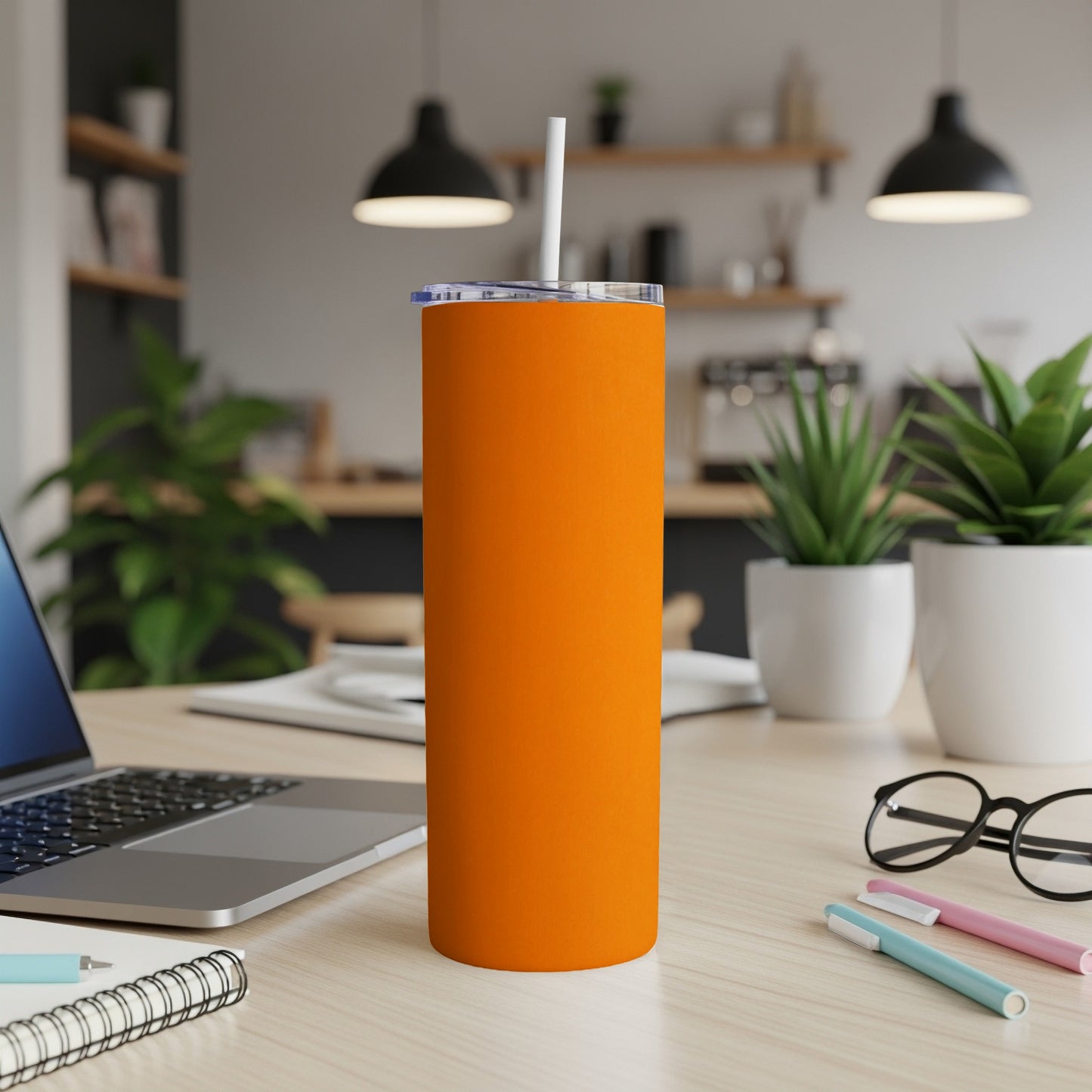 Orange tumbler with straw on a desk next to a laptop and pens.
