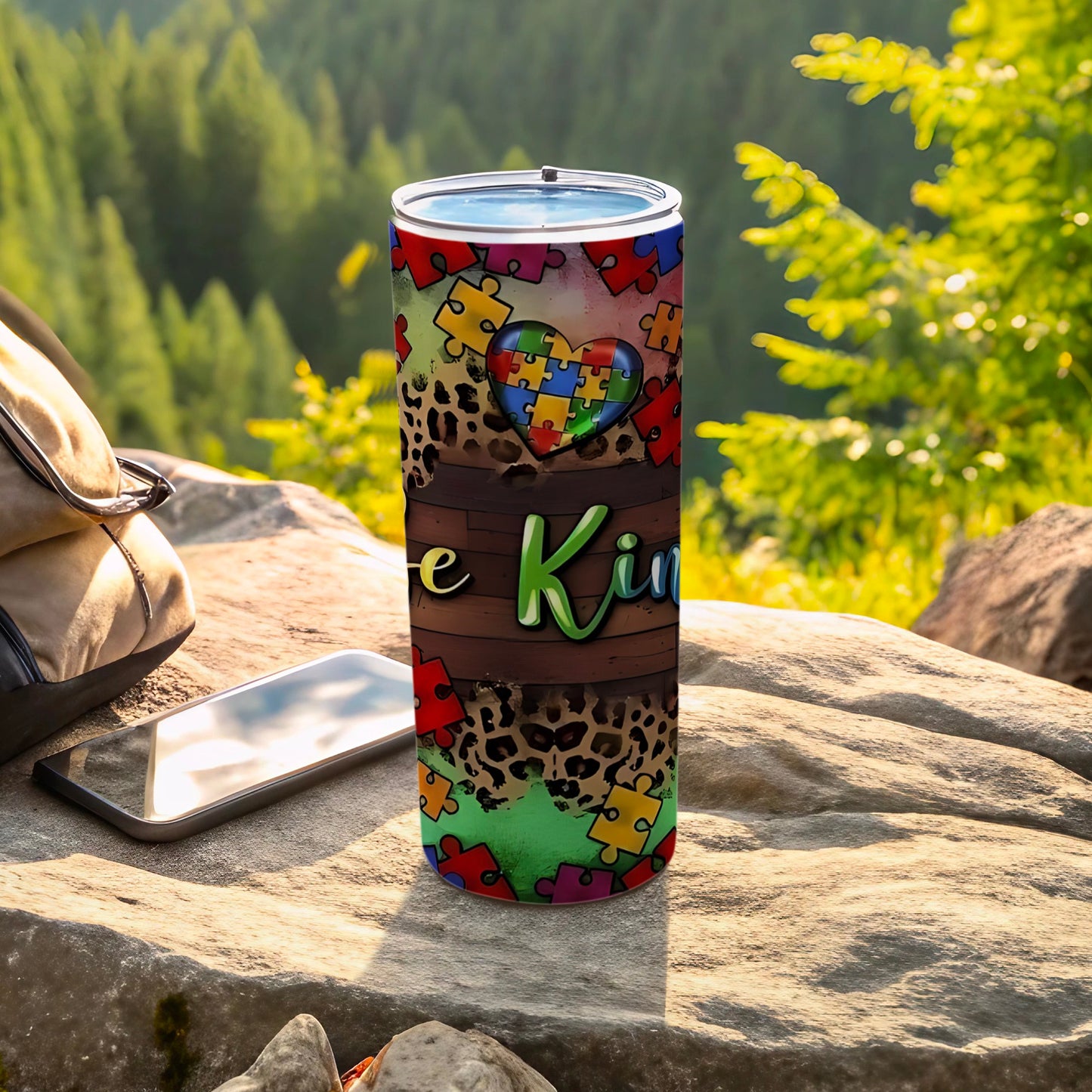 Autism awareness tumbler with puzzle heart and "Be Kind" text on rocky surface.