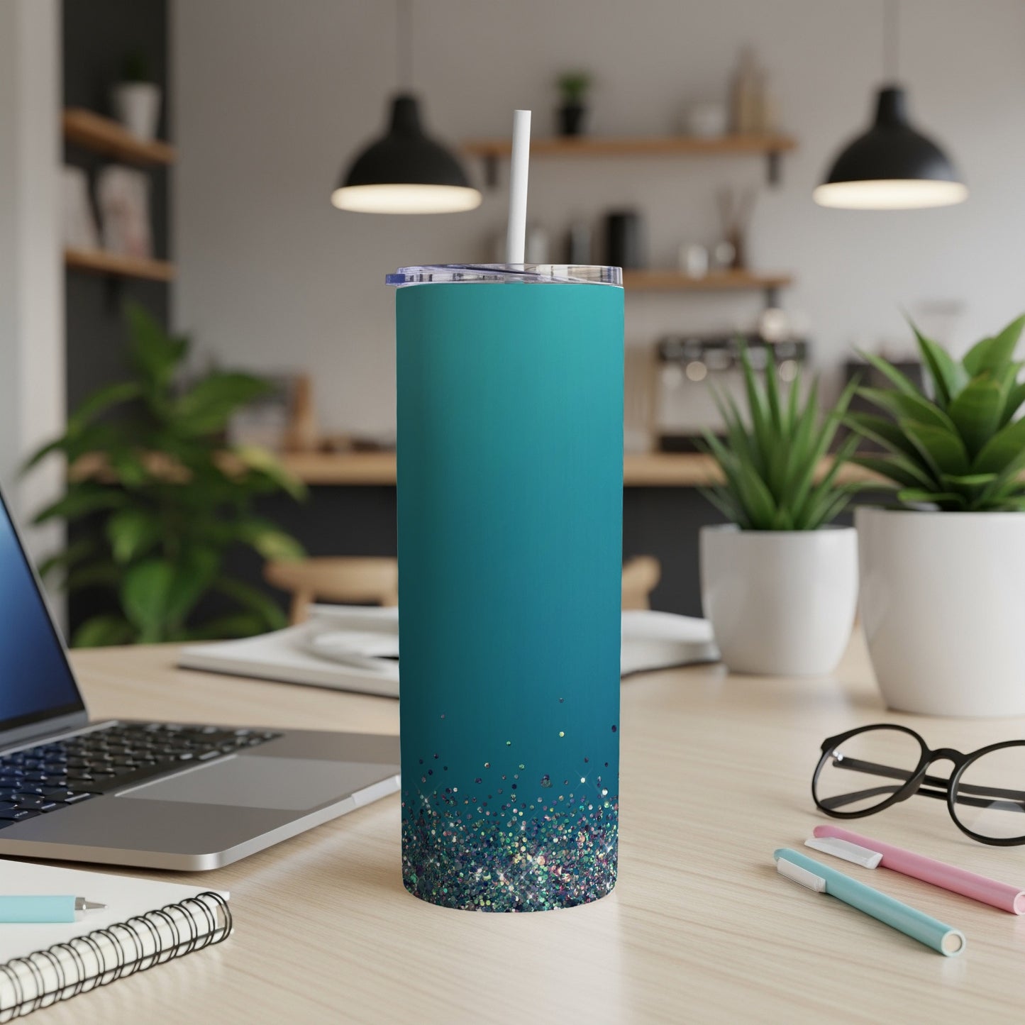 Teal tumbler with glitter bottom and straw on a wooden desk.