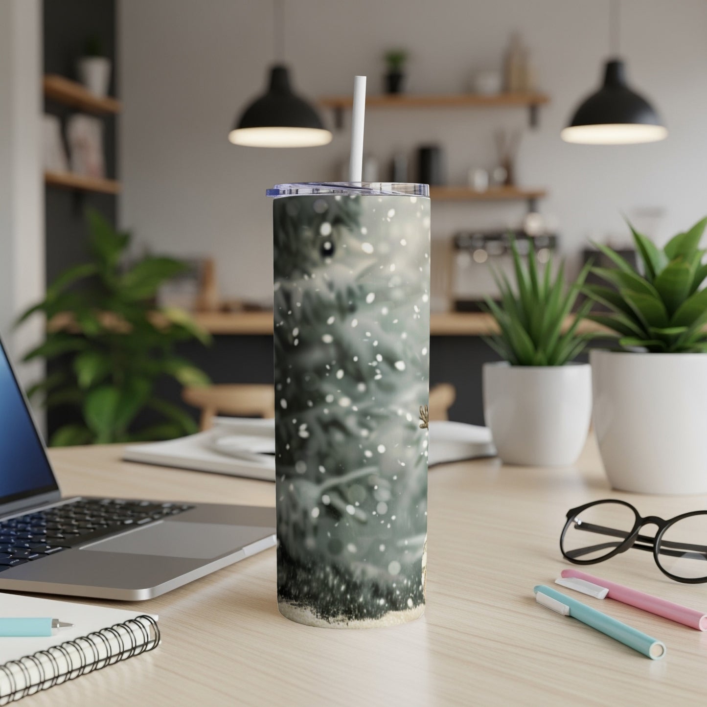 Tumbler with snowy evergreen trees and subtle bokeh, on a desk with a laptop.