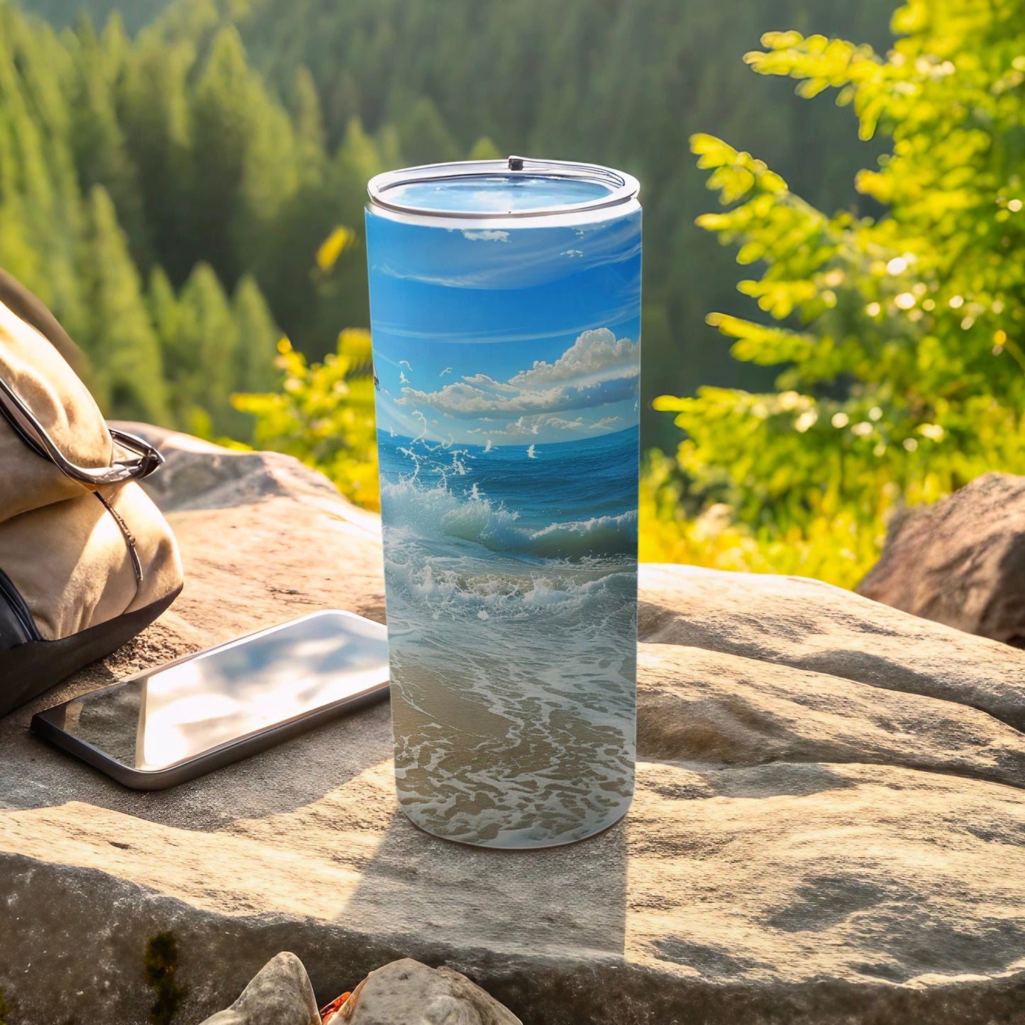 Tall tumbler with ocean wave design sits on rock next to phone and bag.