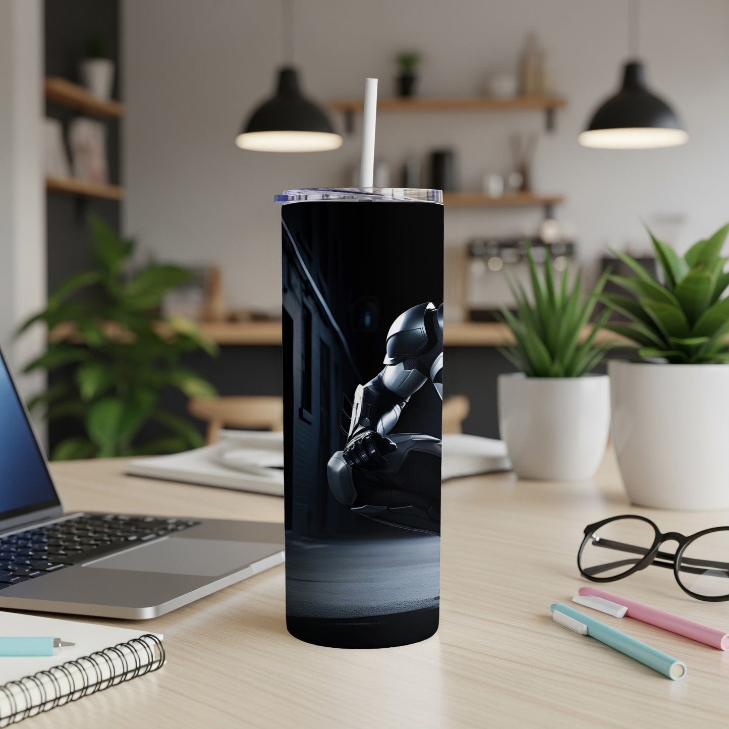 Dark gray tumbler with robot graphic on desk with laptop and pens.