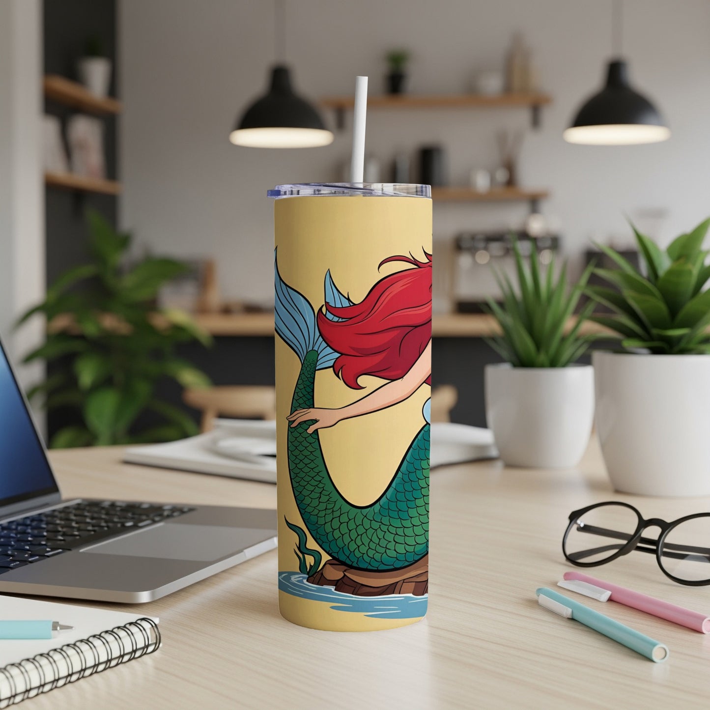 Yellow tumbler with a mermaid illustration, a straw, and office desk accessories.