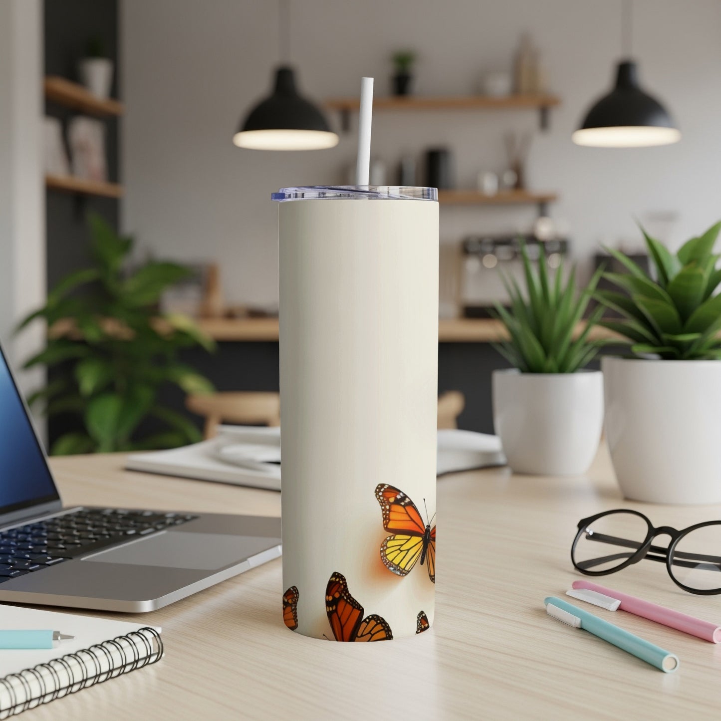 Tall white tumbler with monarch butterflies, next to a laptop and pens.