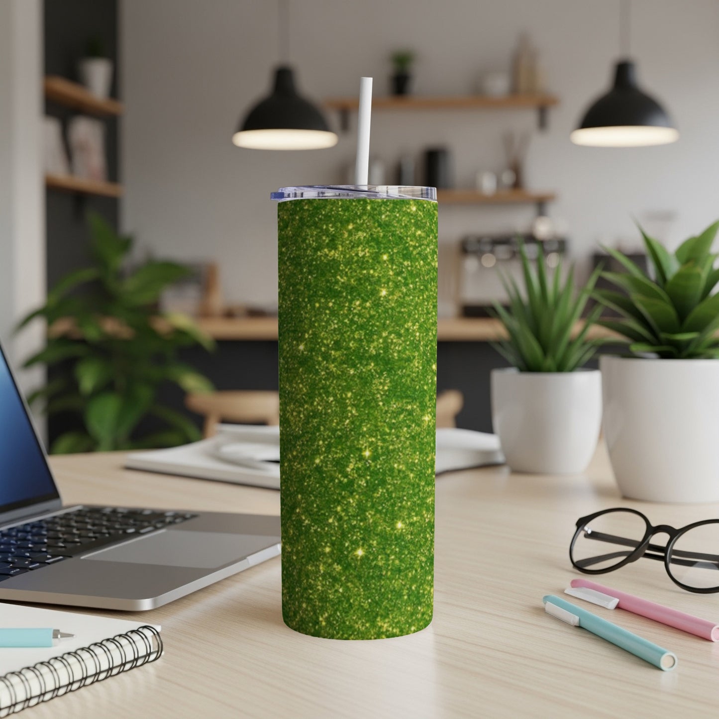 Green glitter tumbler with straw on a desk with laptop and plants.