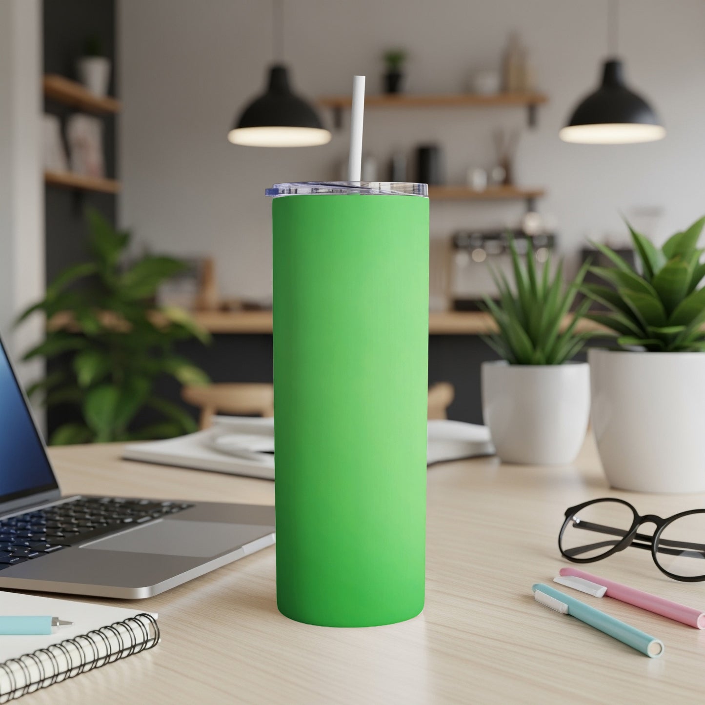Green tumbler with straw on a desk, next to a laptop and pens.