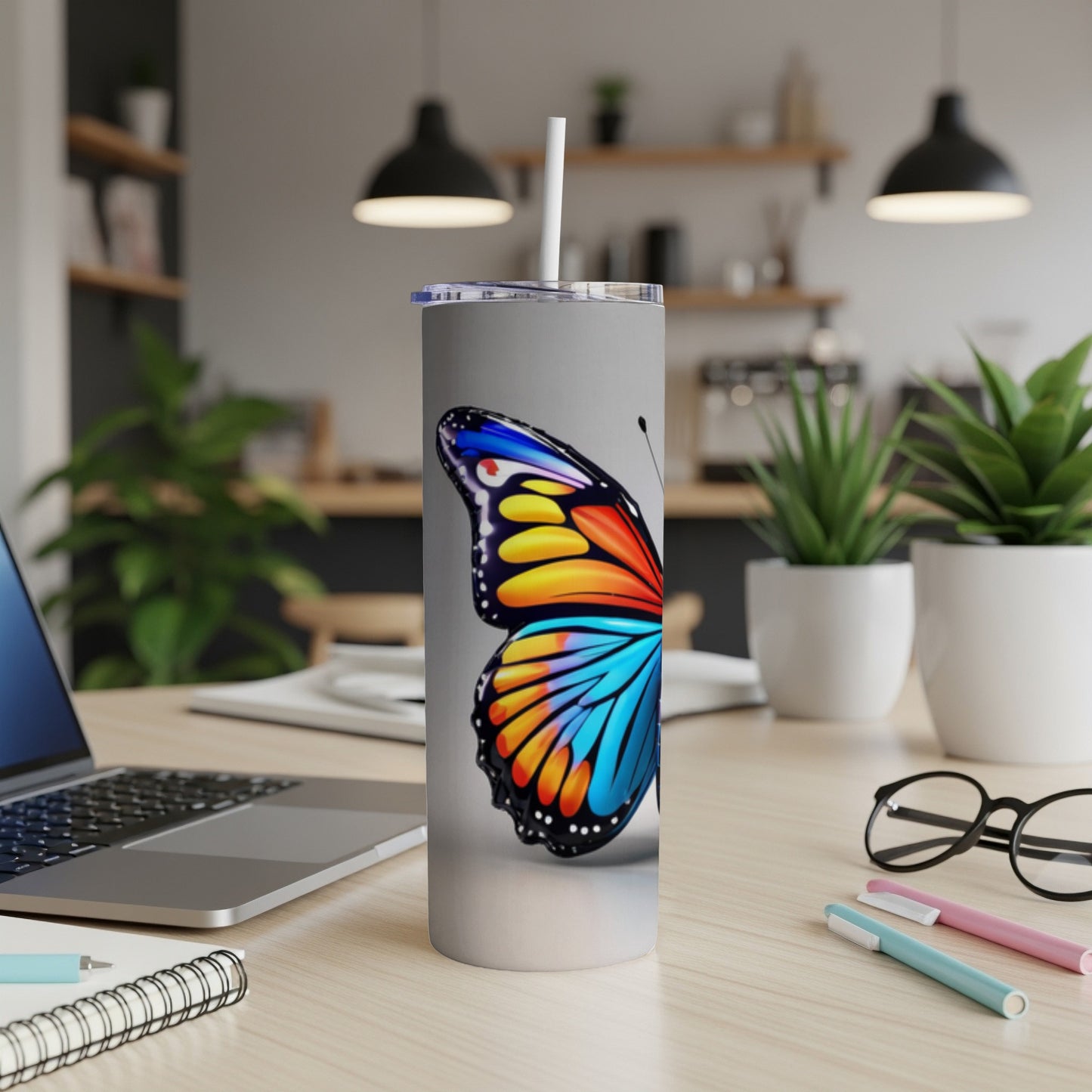 Silver tumbler with colorful butterfly design, straw, on desk with laptop.