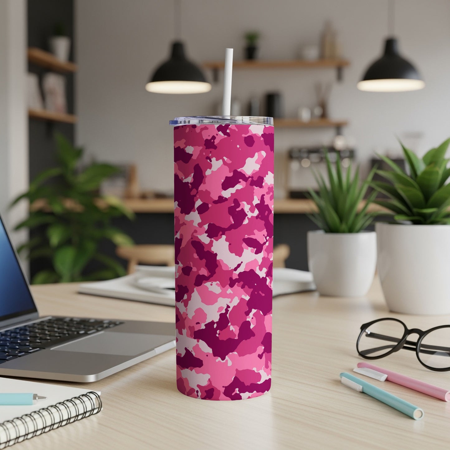 Pink camouflage tumbler with straw on wooden desk next to laptop and notebook.