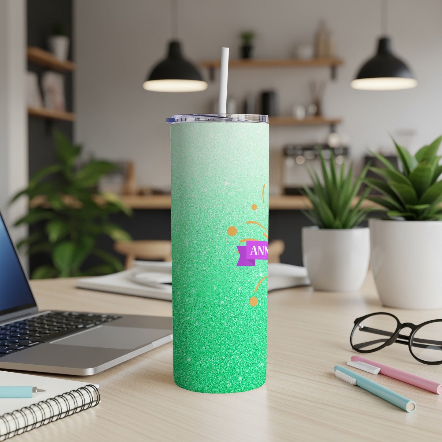 Green glitter tumbler with "ANNIVERSARY" ribbon and straw on desk.