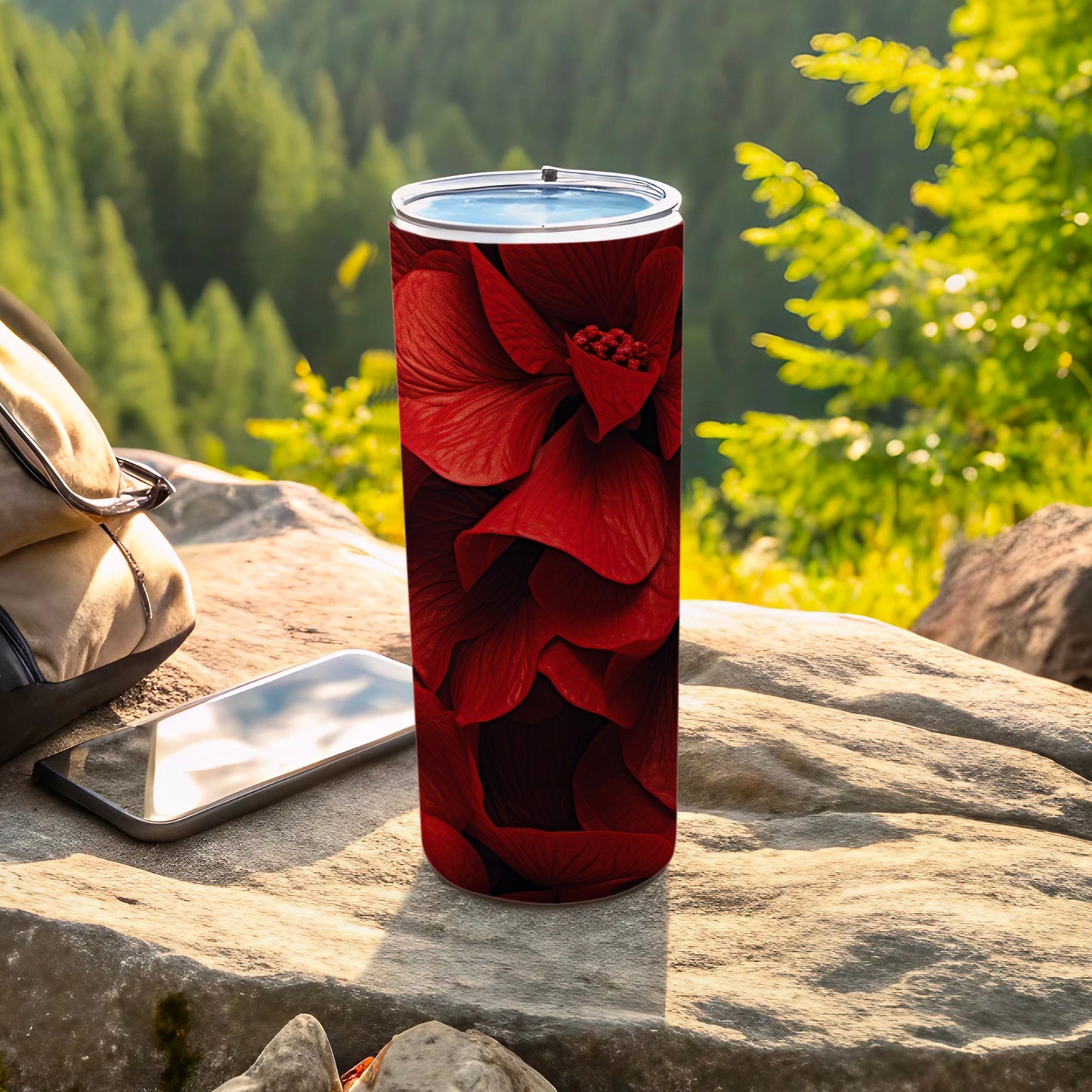 Red floral print tumbler with lid on rock next to phone and backpack.