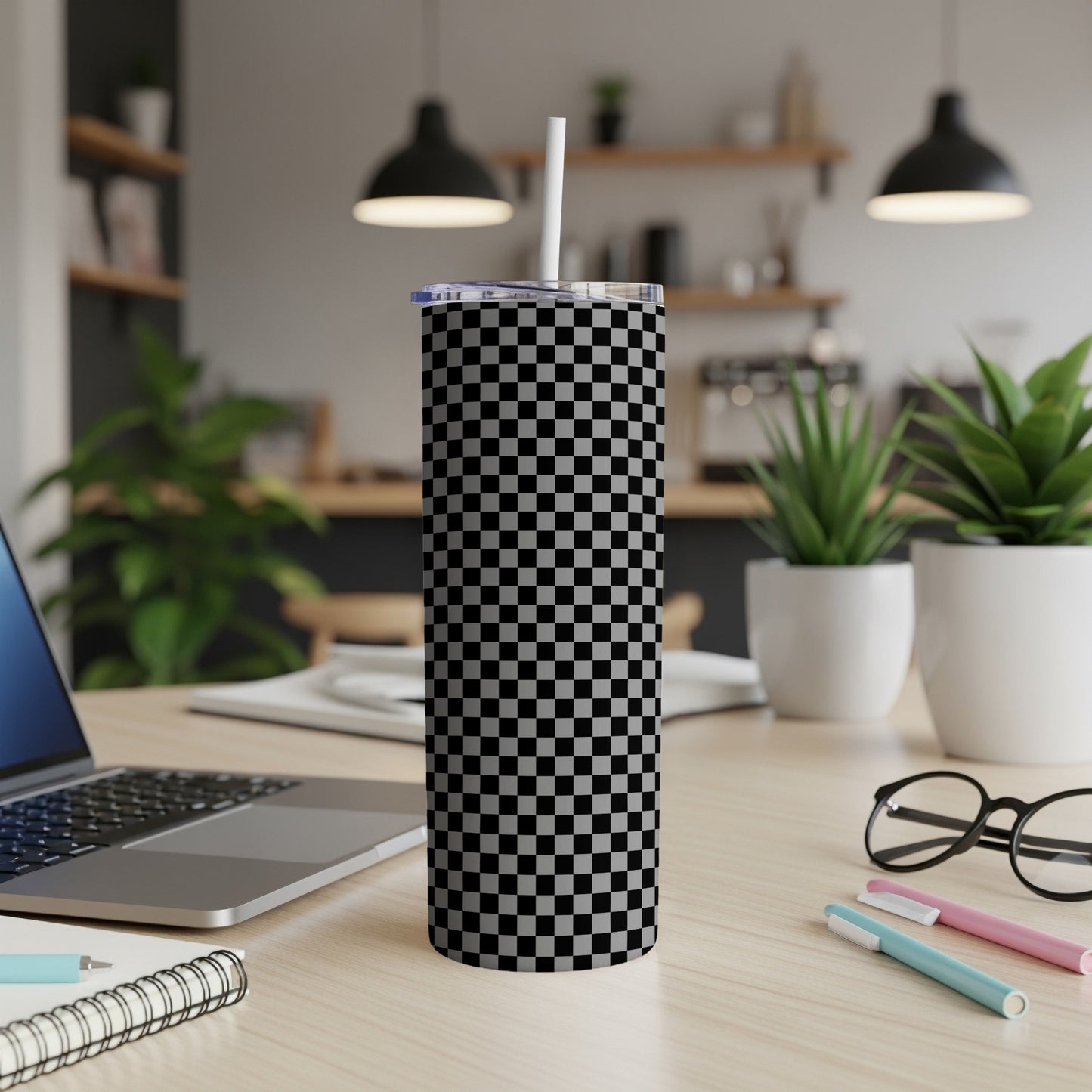 Checkered tumbler with straw on a desk with a laptop and plants.