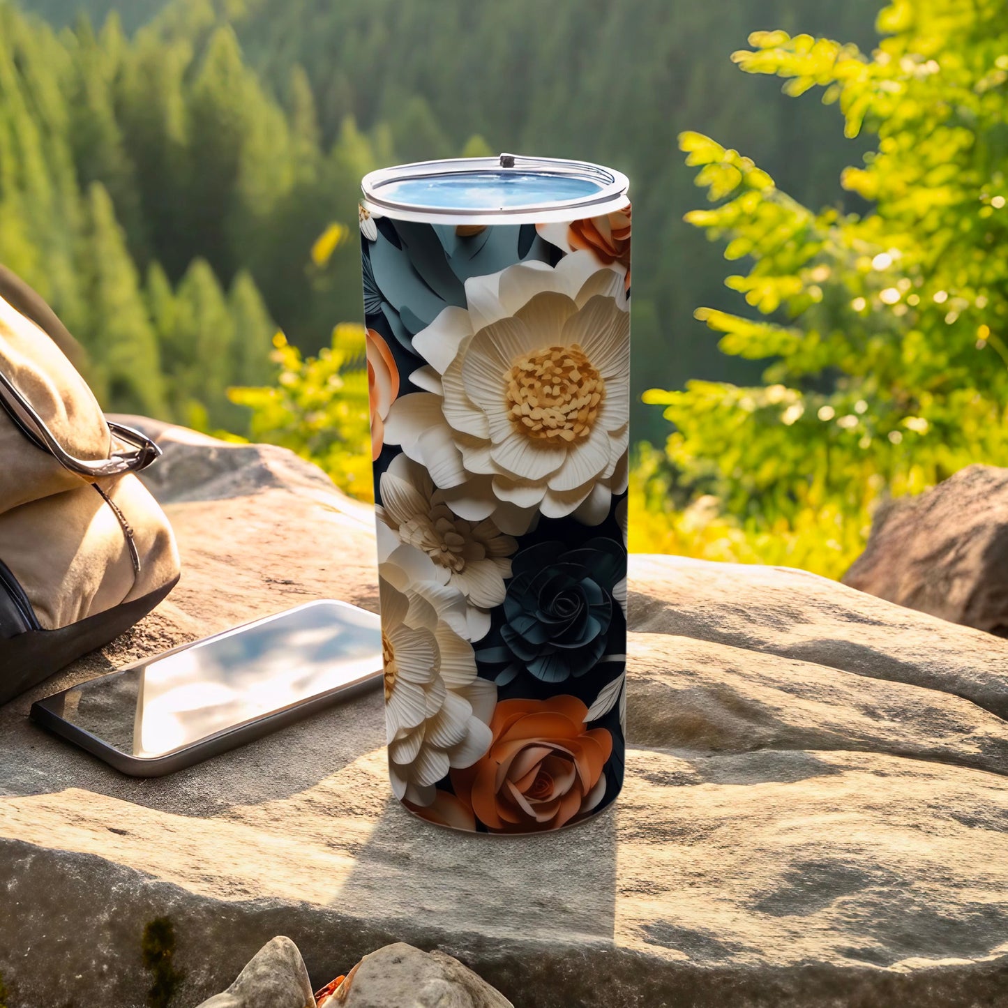 Tall tumbler with floral design on a rock with a phone and backpack.