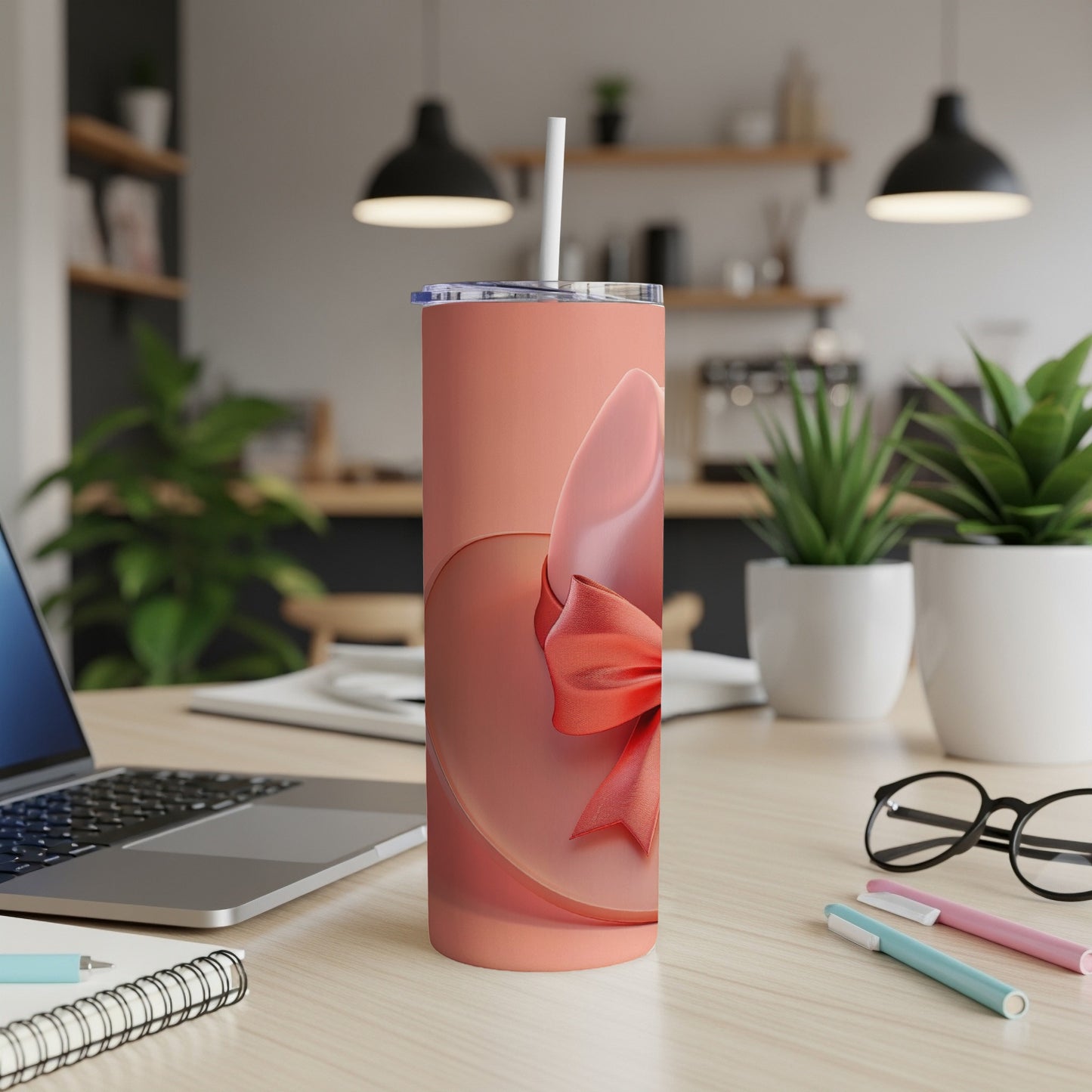 Coral tumbler with a red ribbon accent sits on a desk with a laptop and pens.