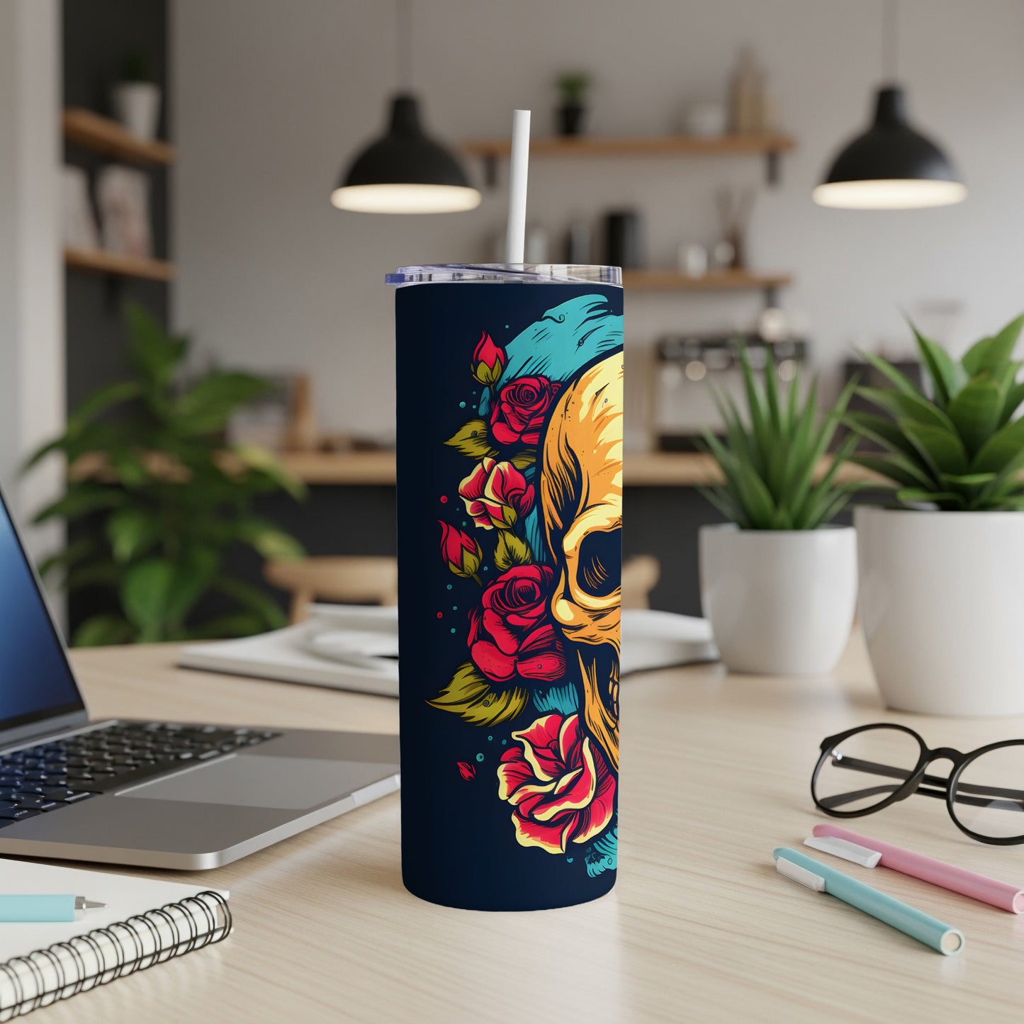 Dark tumbler with skull and roses, on a desk with laptop and pens.