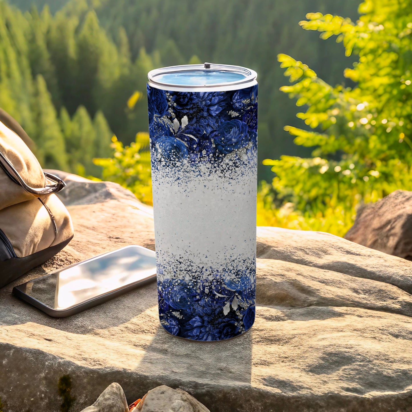 Blue floral tumbler with glitter-like pattern on a rock with phone and bag.