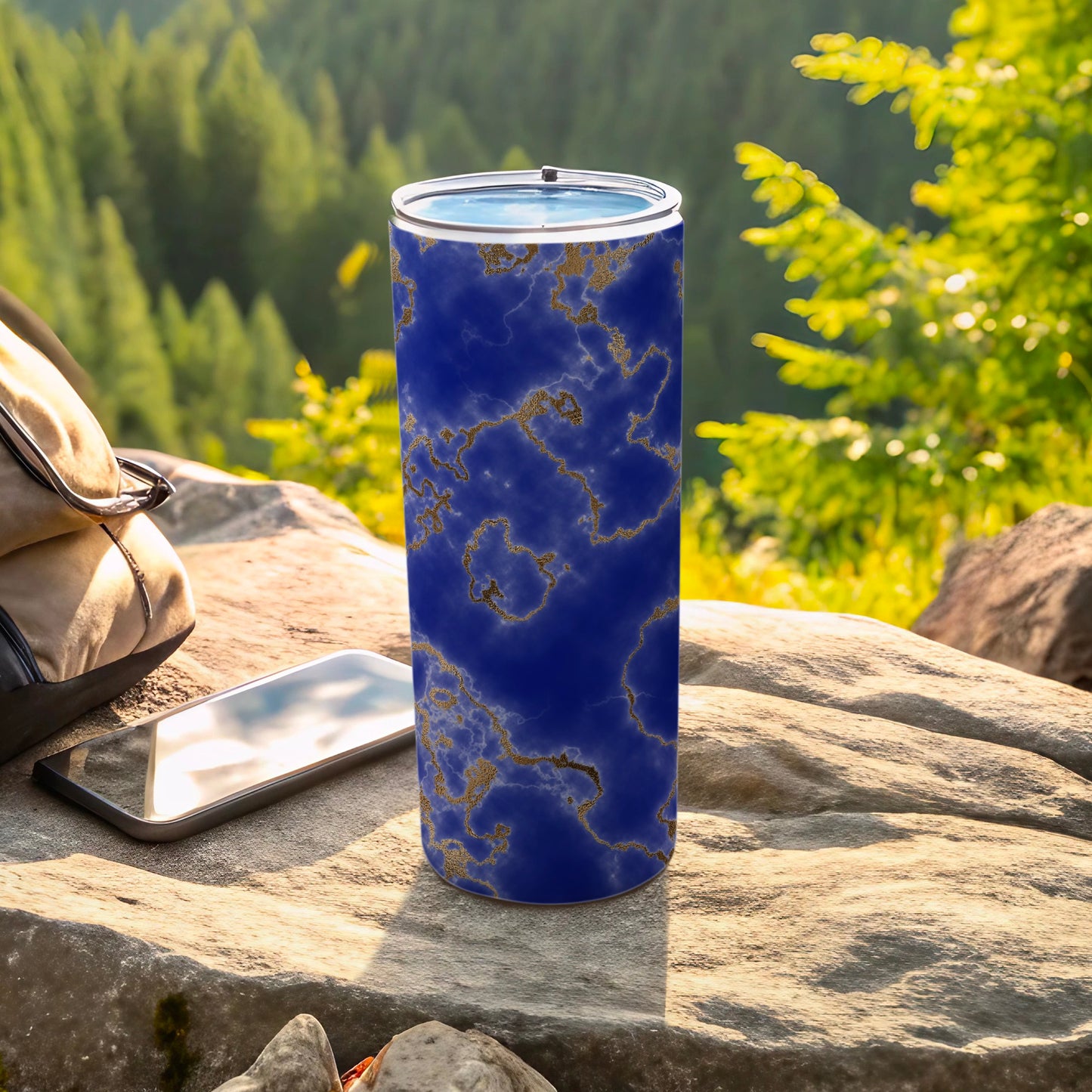 Blue and gold marble pattern tumbler on rock with phone and bag.