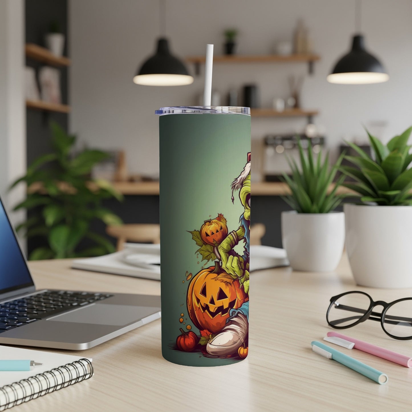 Green tumbler with cartoon zombie and pumpkins, on a desk with laptop and glasses.