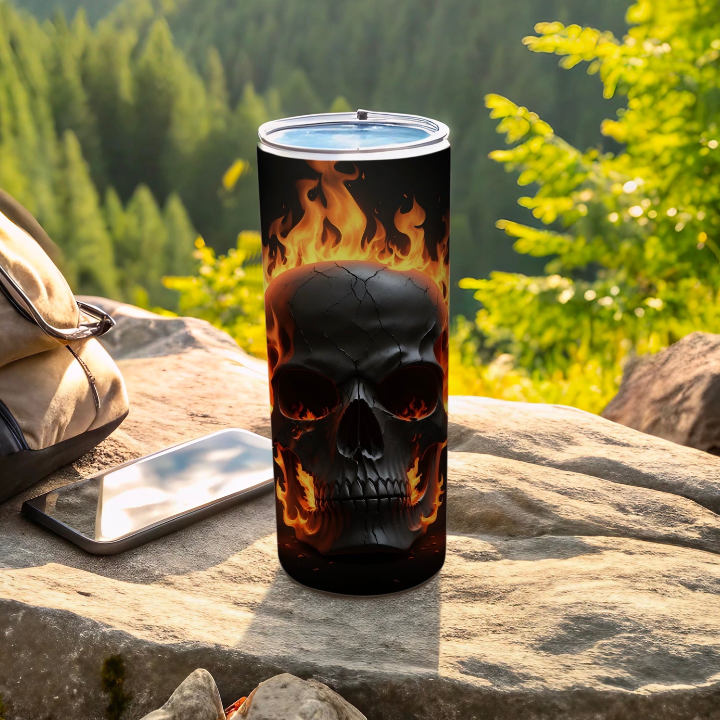 Black tumbler with flaming skull design sits on rock with backpack and phone.
