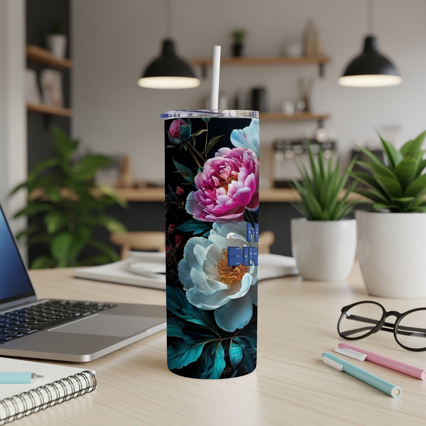 Floral tumbler with a straw on a desk with a laptop and pens.