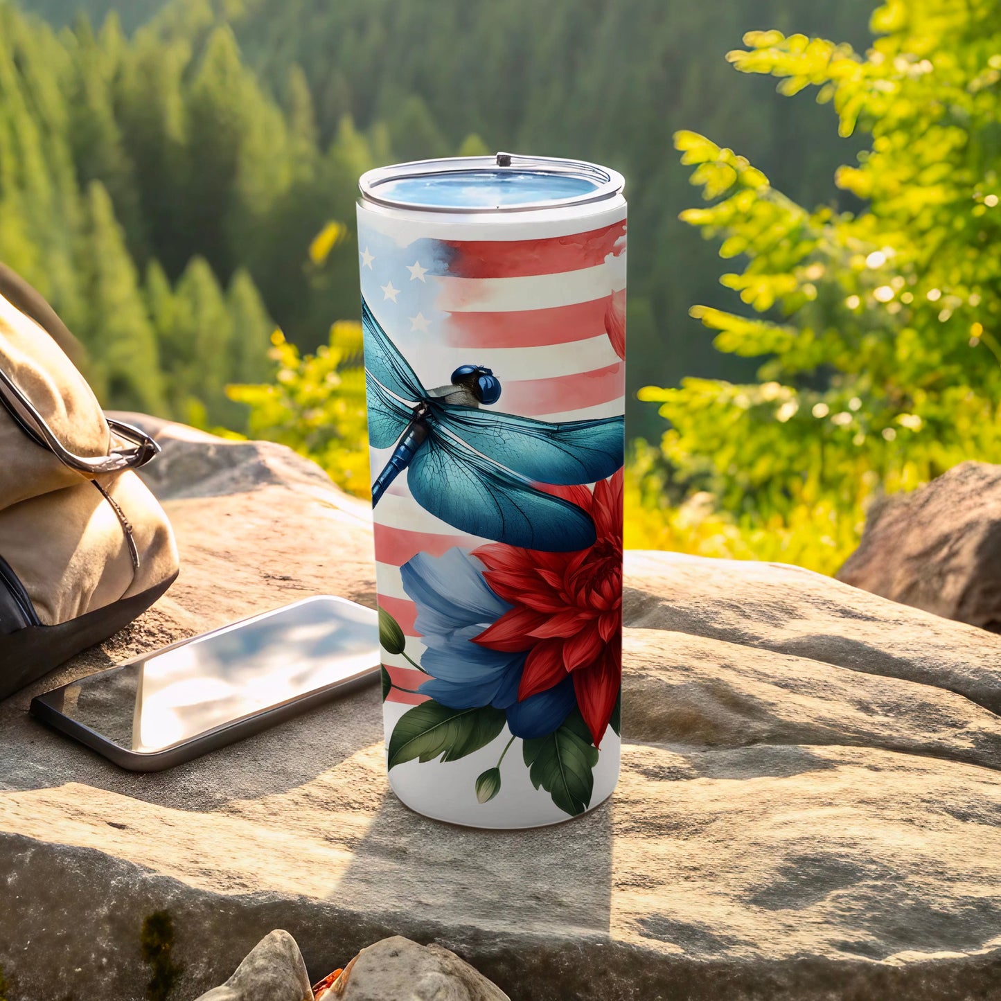 White tumbler with American flag, dragonfly, and floral design, on rocky surface outdoors.