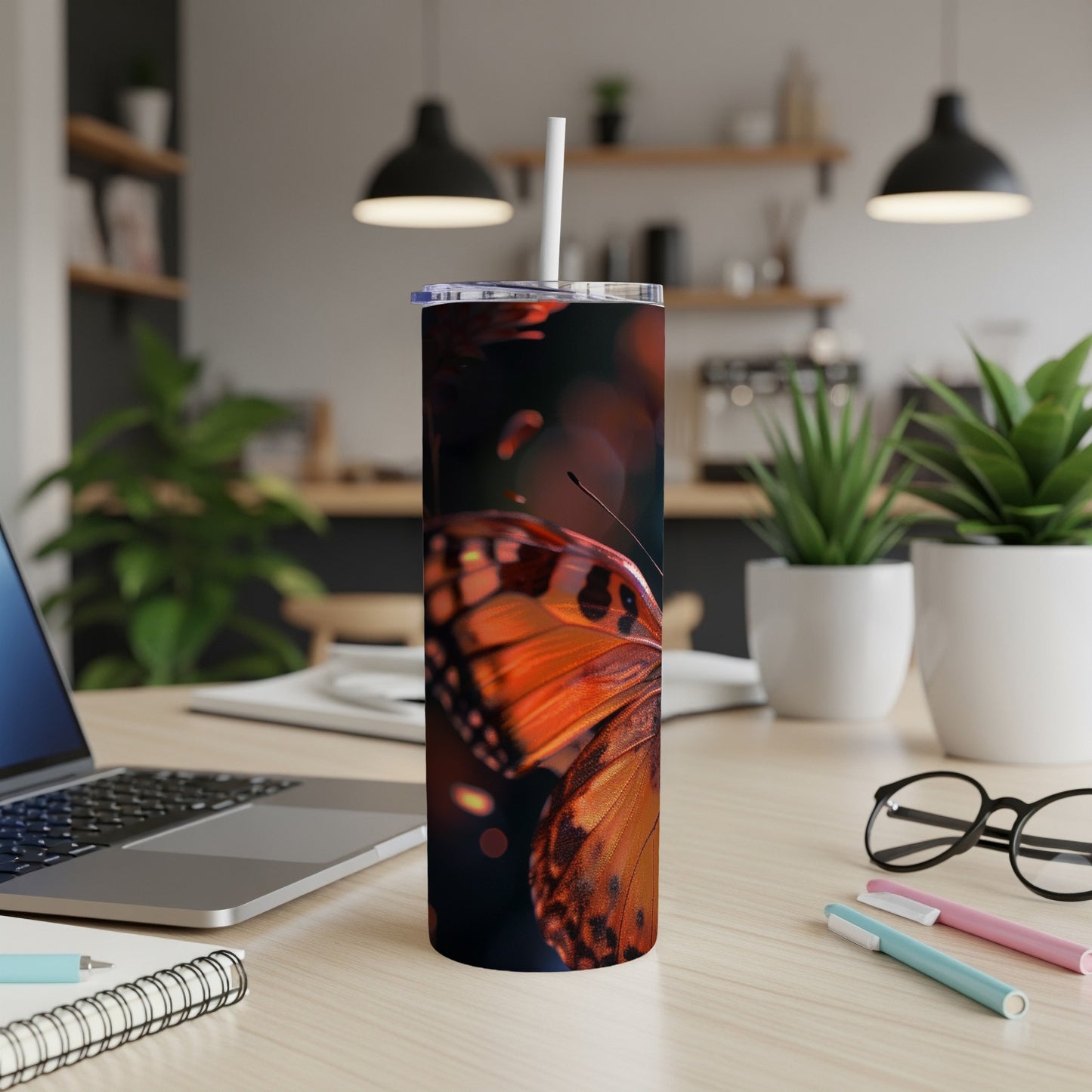 Tumbler with orange butterfly wings, desk with laptop, plants, and pens.