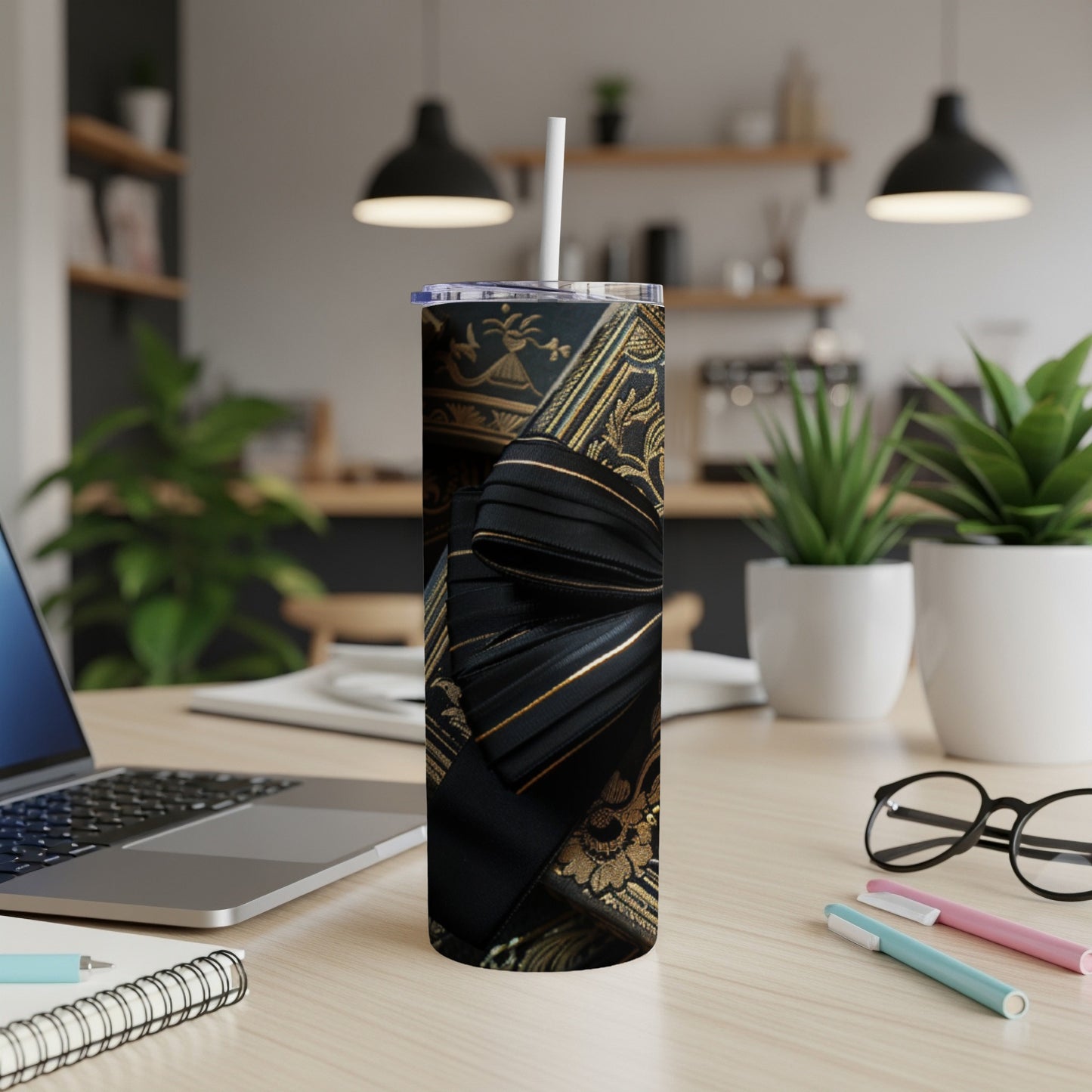 Black and gold patterned tumbler with ribbon design, straw, and office desk items.