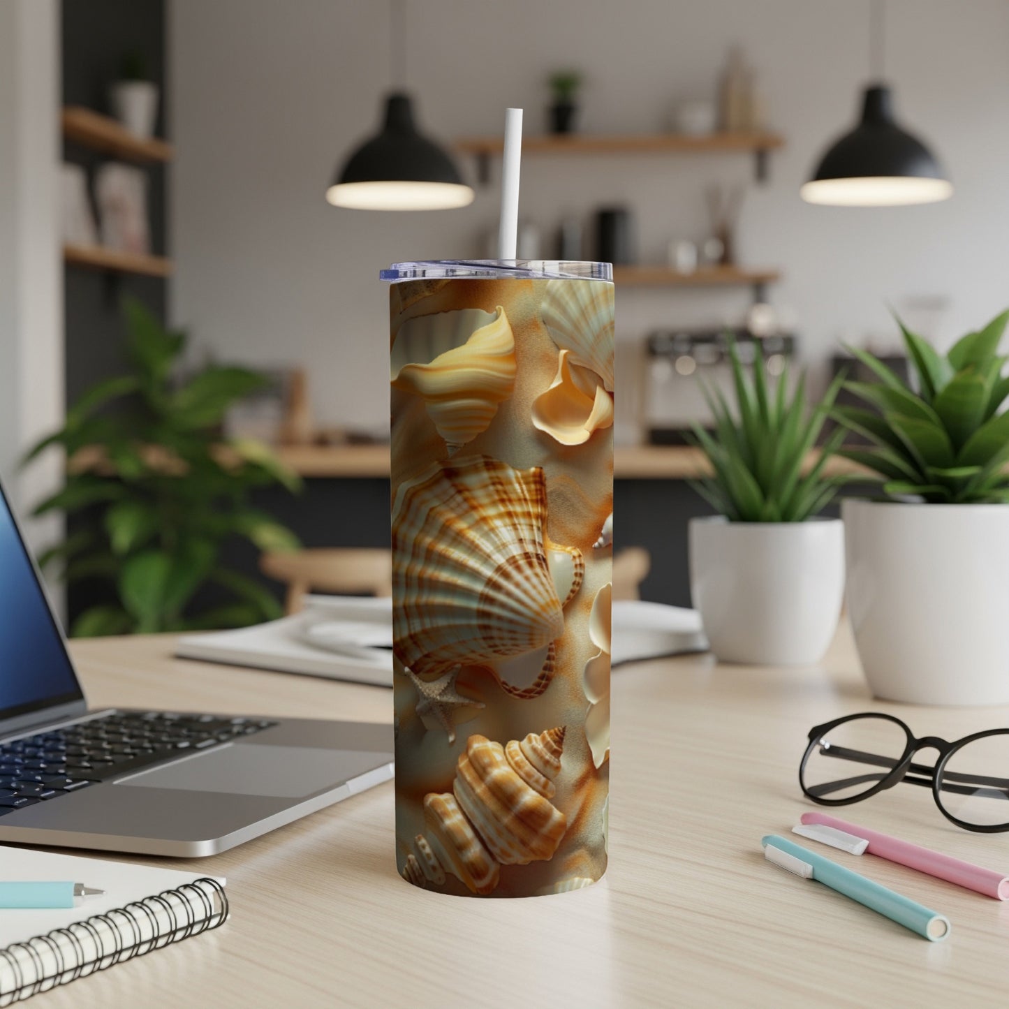 Tall tumbler with seashell design on a desk with laptop and pens.