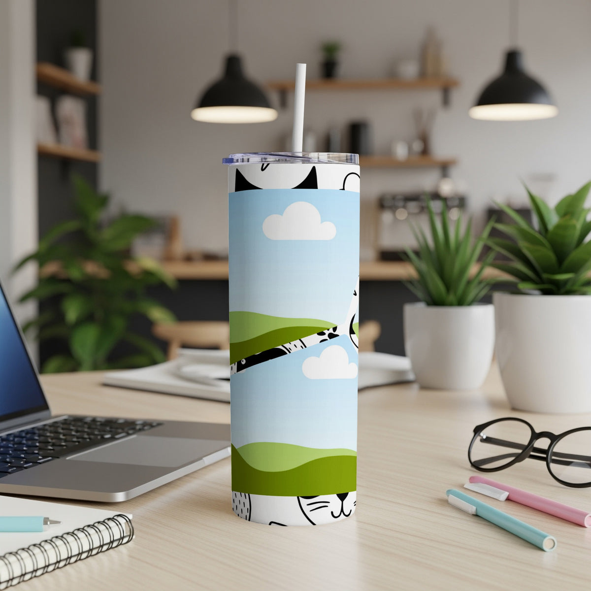 Tumbler with playful cat and cloud design on a desk with a laptop and pens.