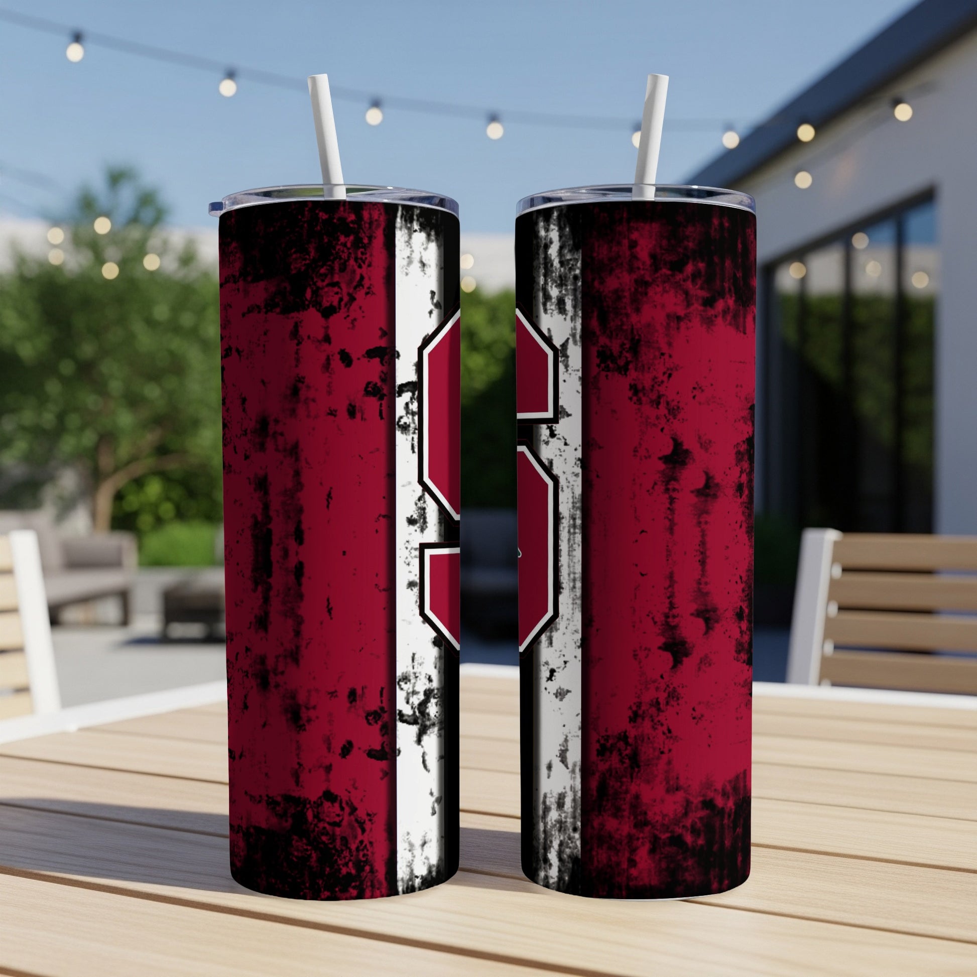 Two red and white distressed tumblers with straws sit on a wooden table outside.