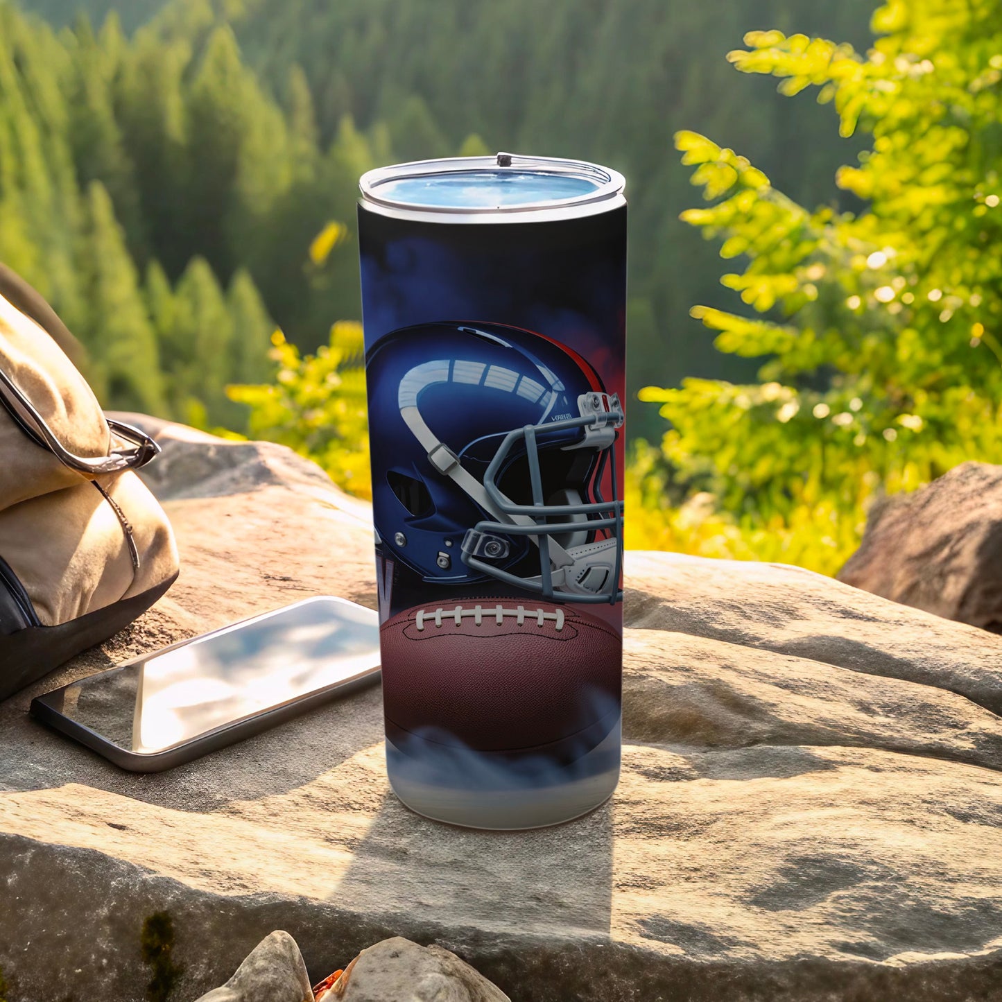 Tumbler with football helmet and ball graphic, on rocks with backpack and phone.