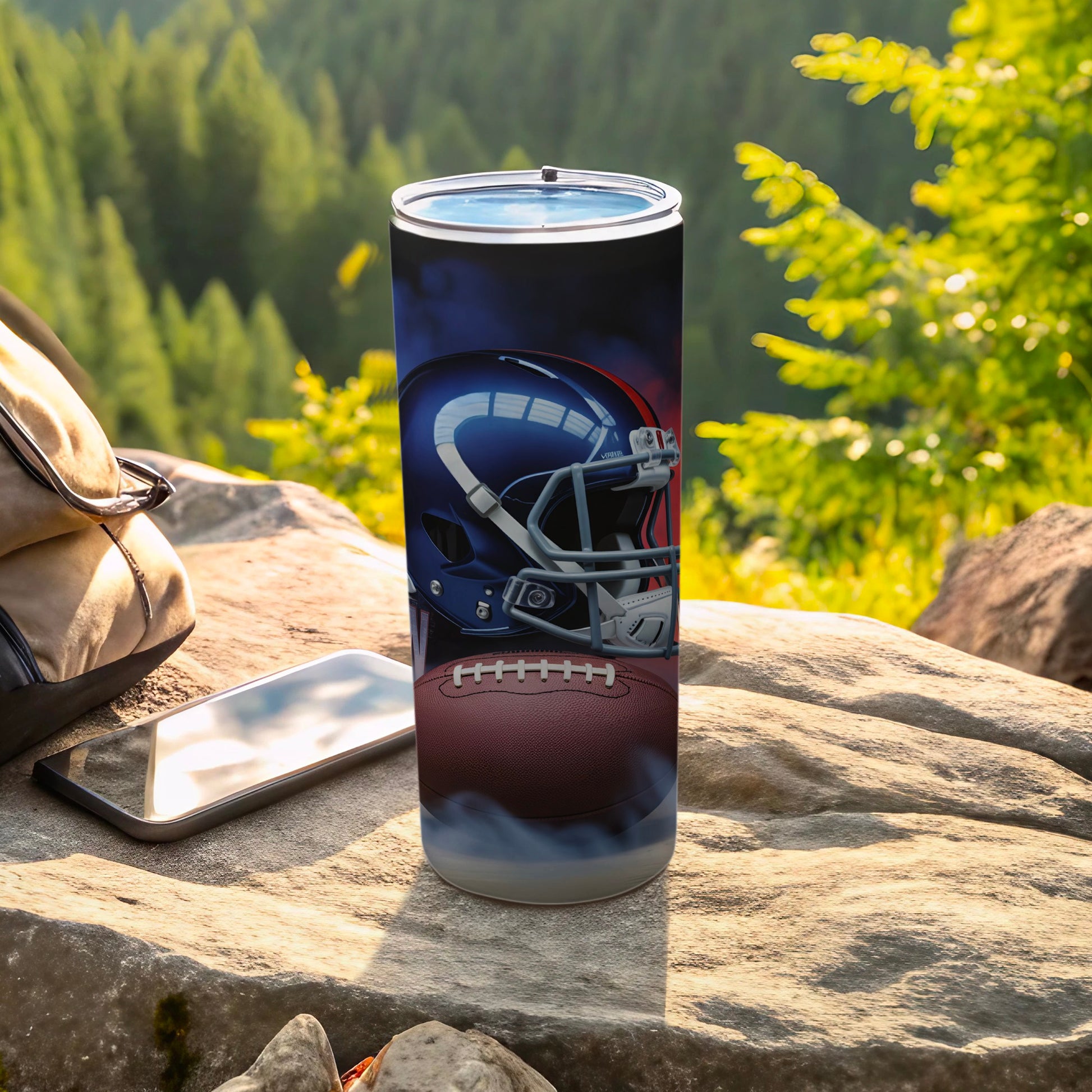Tumbler with football helmet and ball graphic, on rocks with backpack and phone.