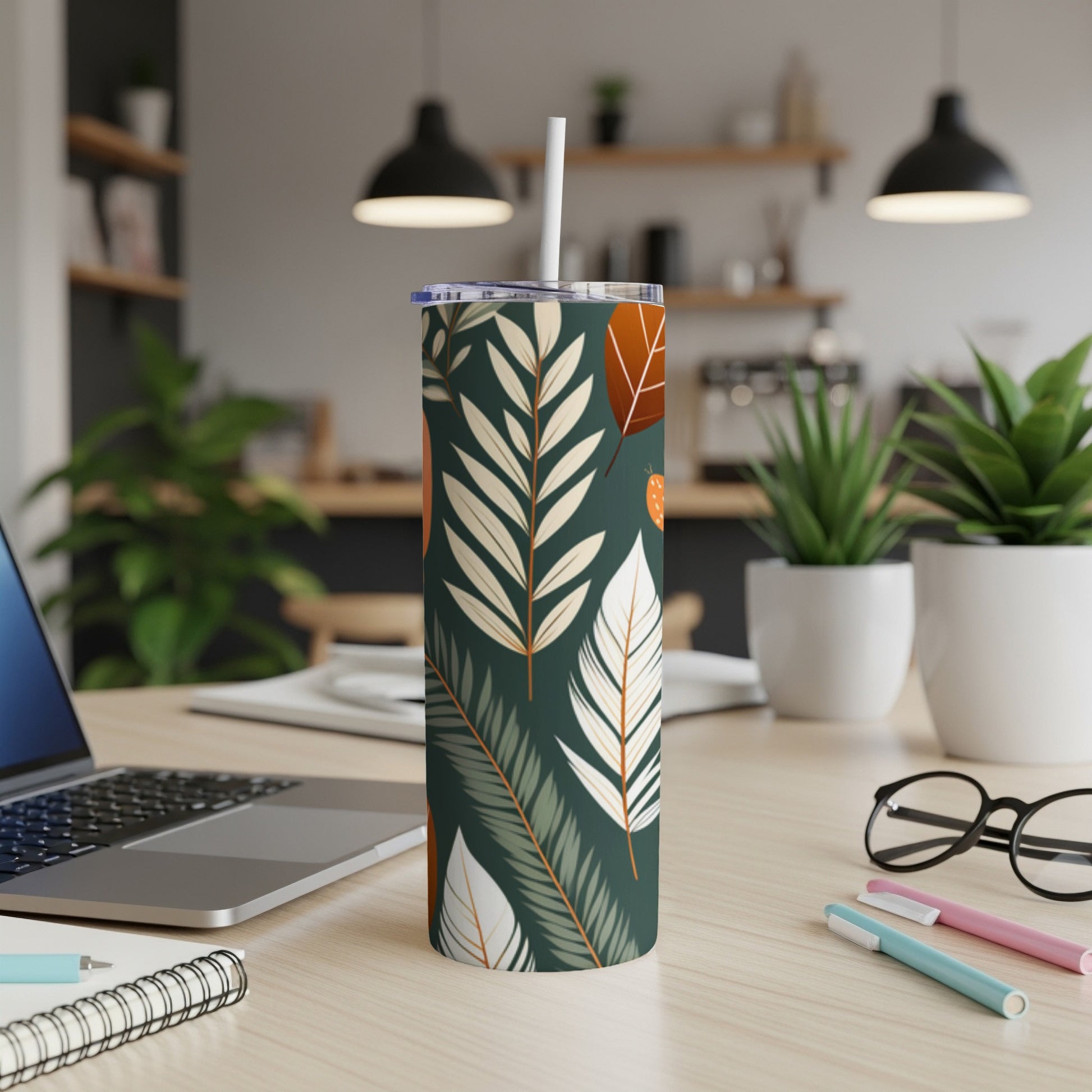 Tumbler with botanical leaf pattern, white straw, laptop, and pens on desk.