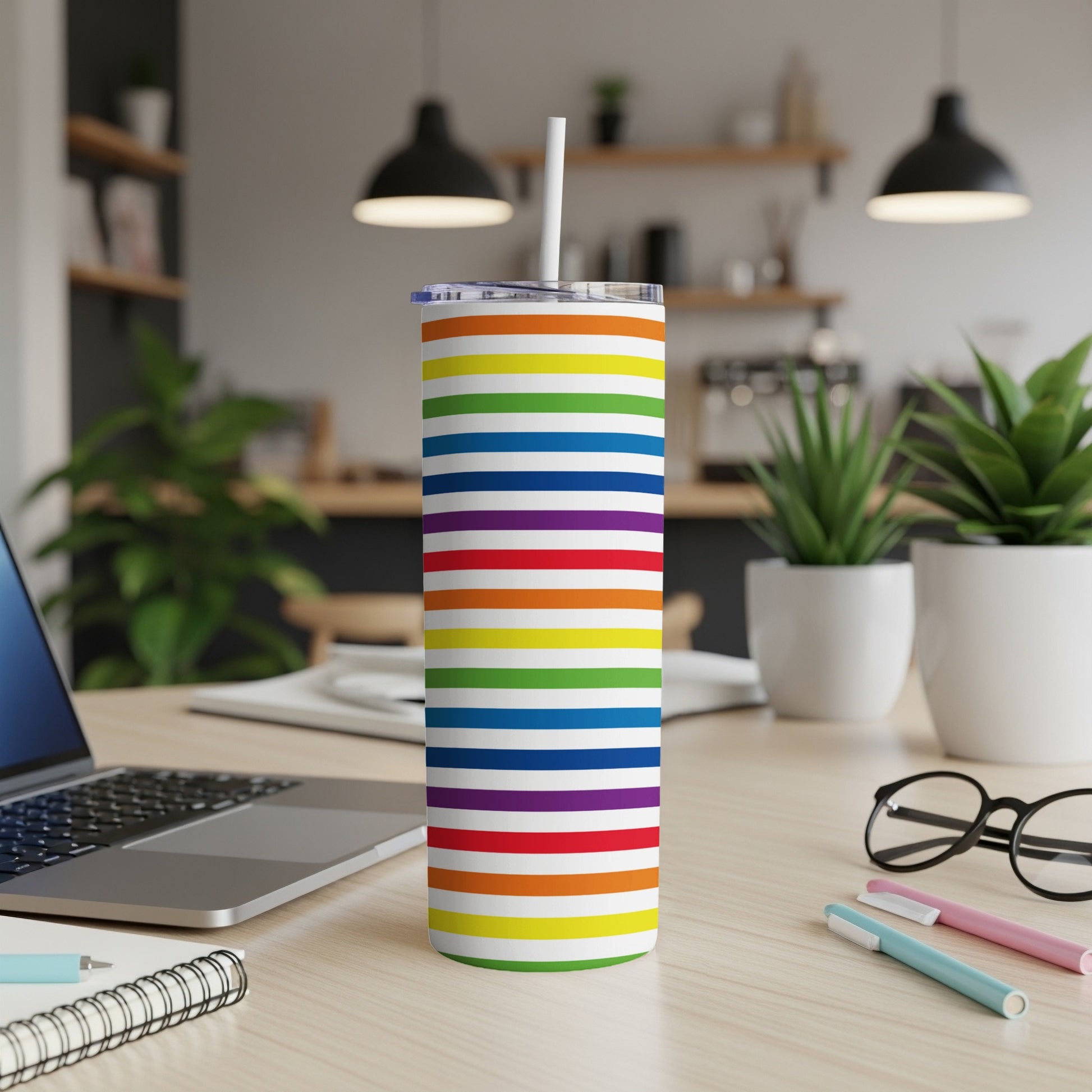 Rainbow striped tumbler with white straw on a desk with a laptop and pens.