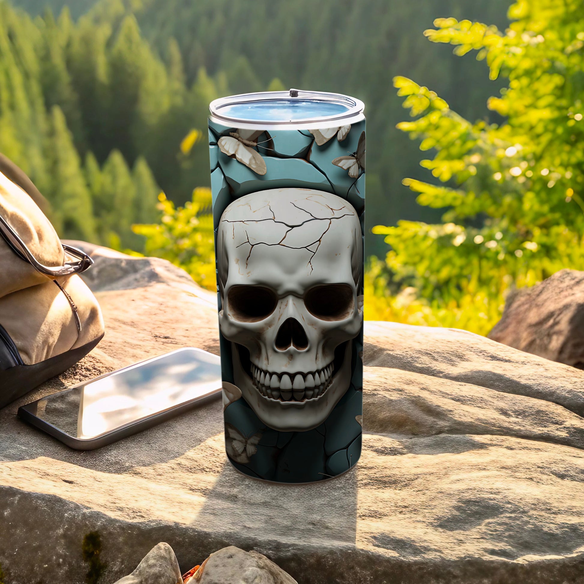 Skull and butterflies tumbler on rock next to backpack and phone.