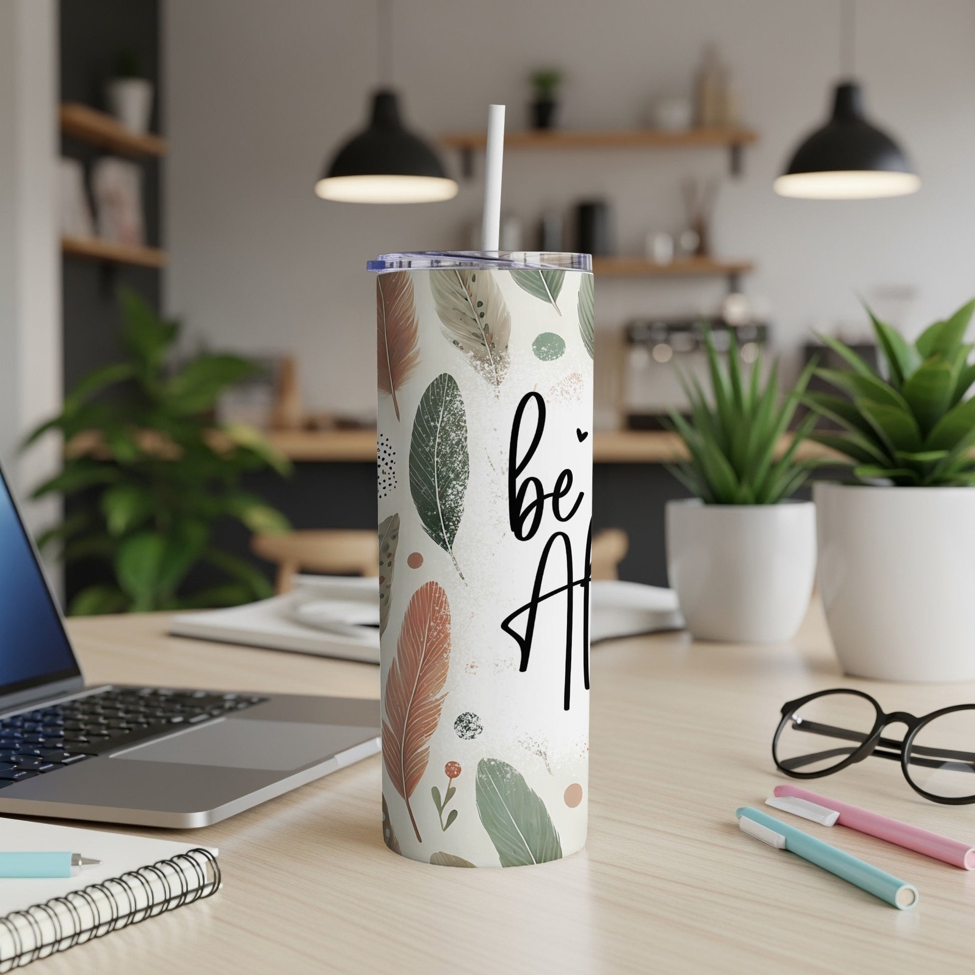 Tall tumbler with feather pattern and "be kind" text, on a desk.