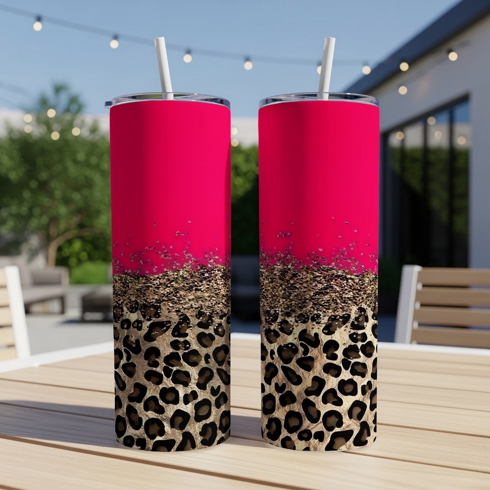 Two pink and leopard print tumblers with straws on a table outdoors.