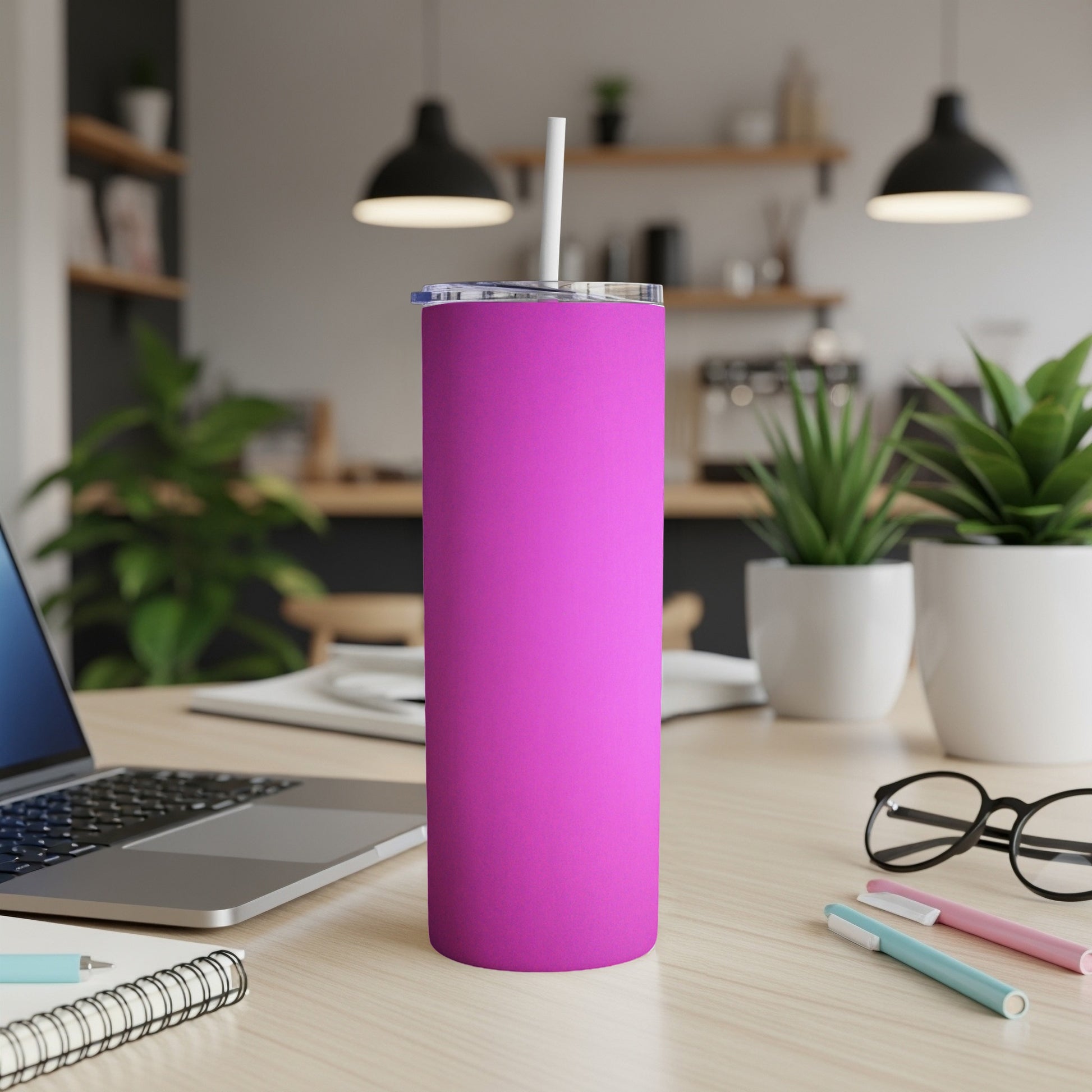 Pink tumbler with white straw on a desk with a laptop and pens.