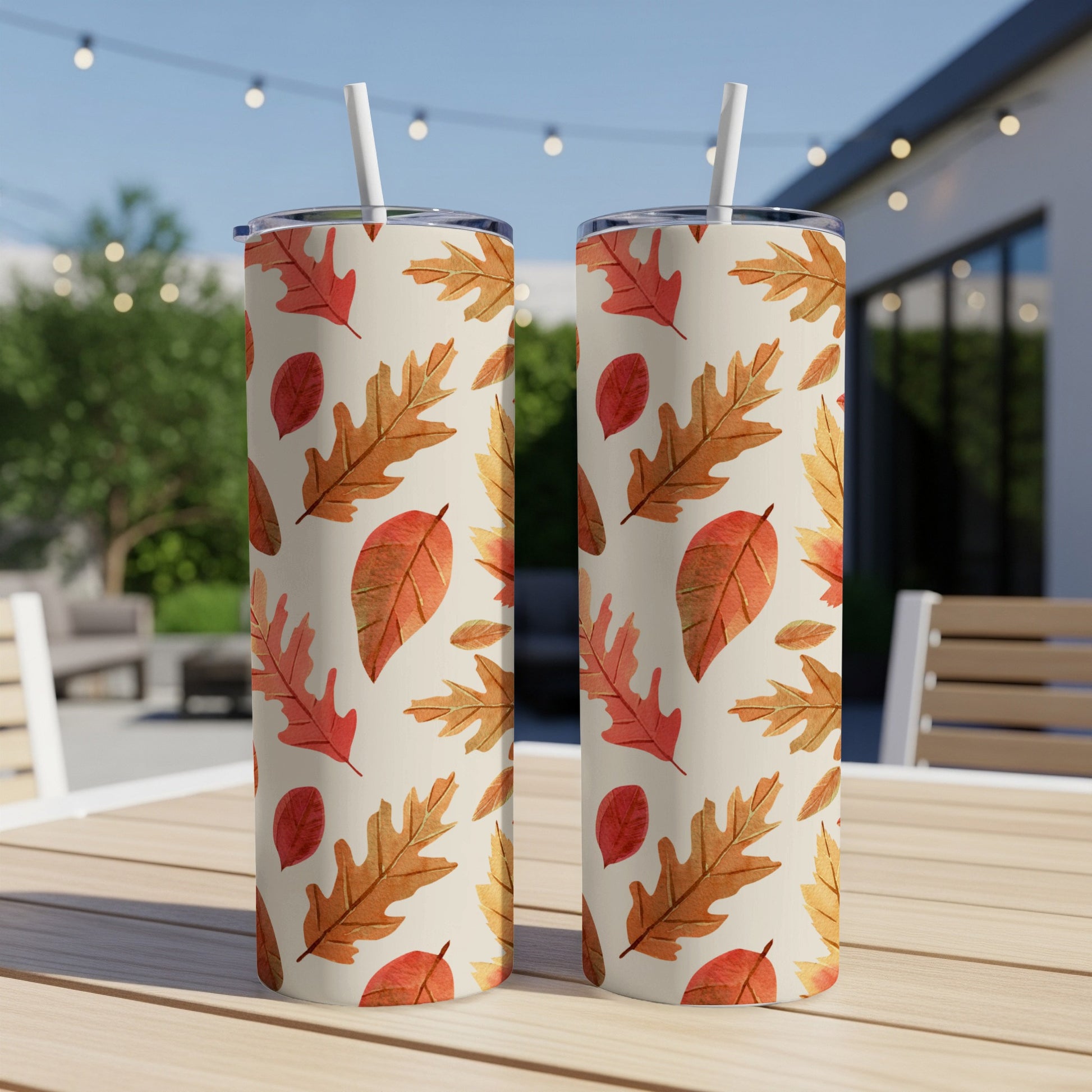 Two tall tumblers with a repeating pattern of autumn leaves on an outdoor table.