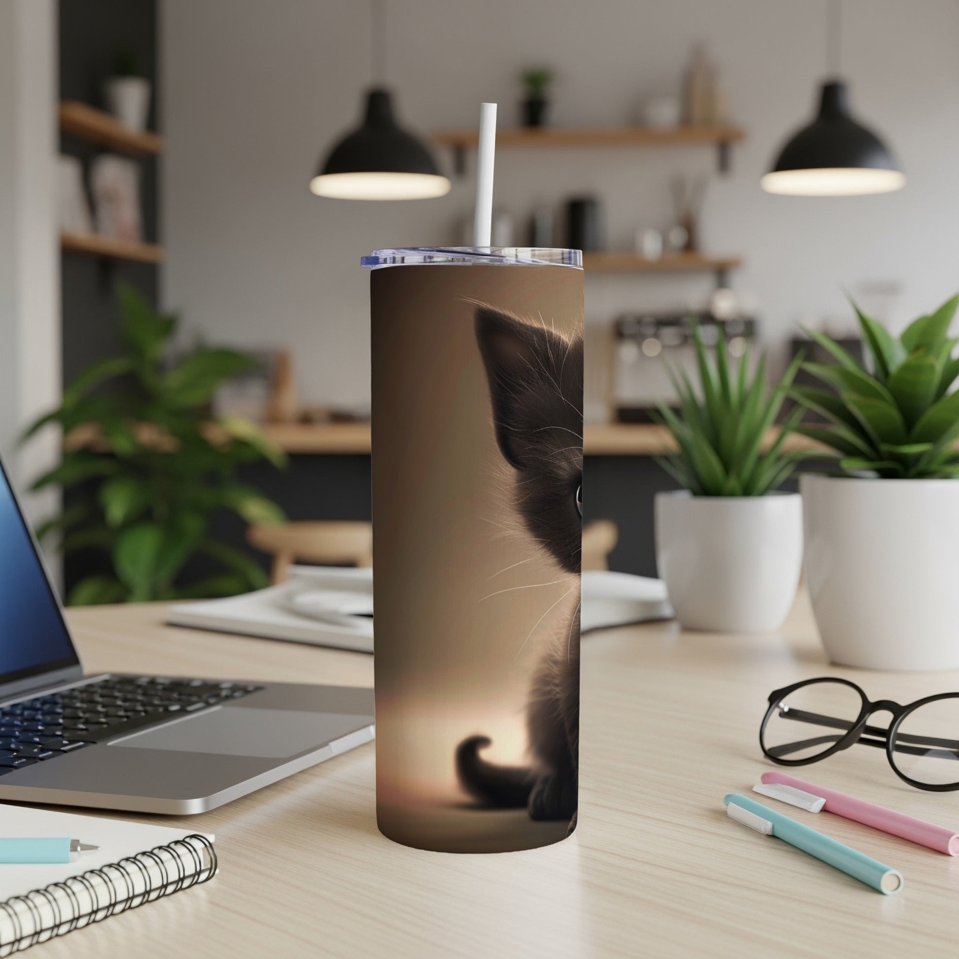 Black kitten peeking from a tall, decorated tumbler on a desk.