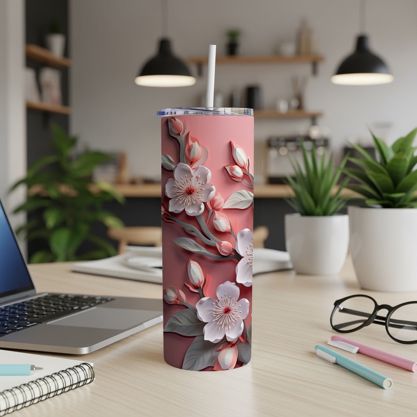 Pink floral tumbler with straw on a desk, next to a laptop and pens.