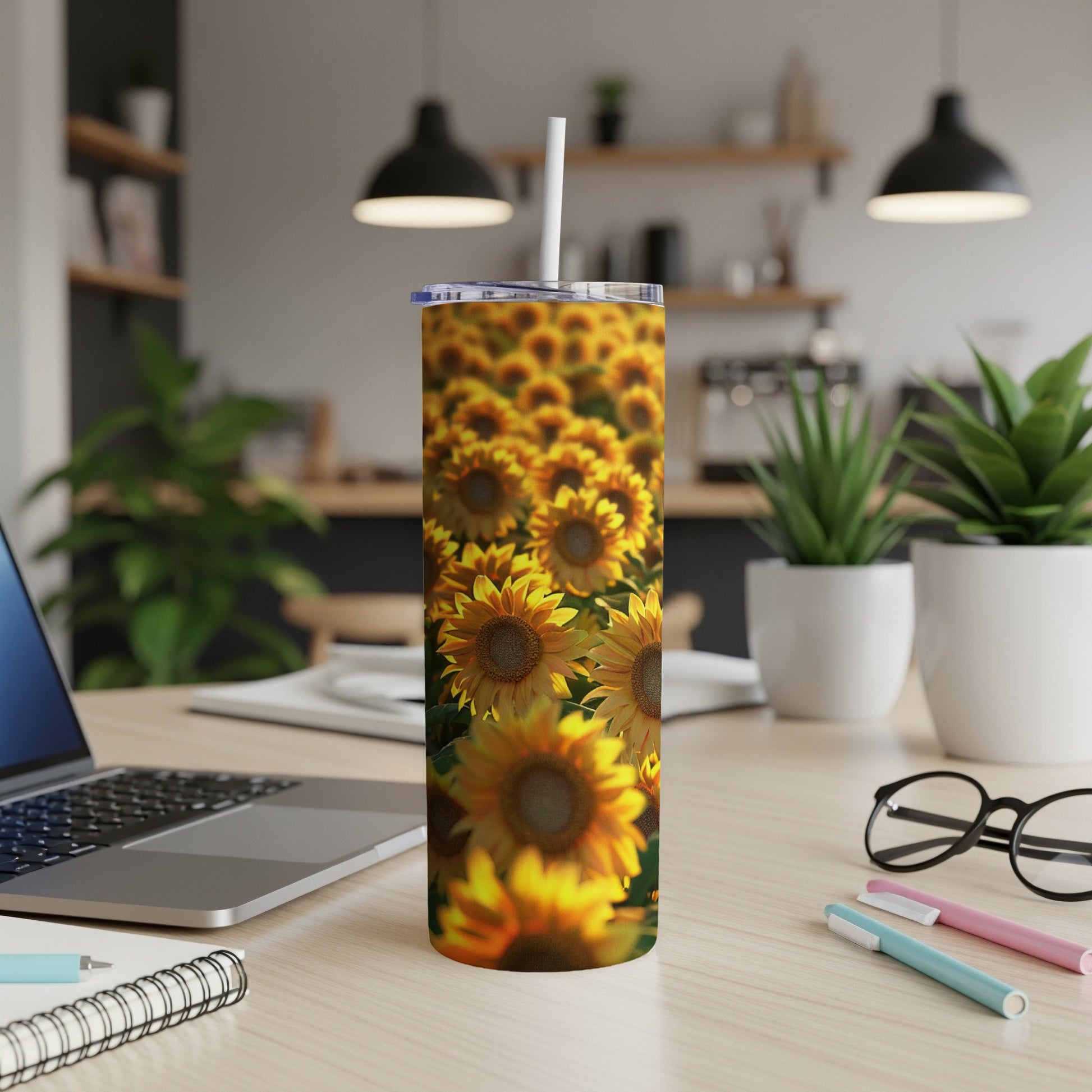 Tall tumbler with sunflowers, white straw, on desk with laptop and pens.