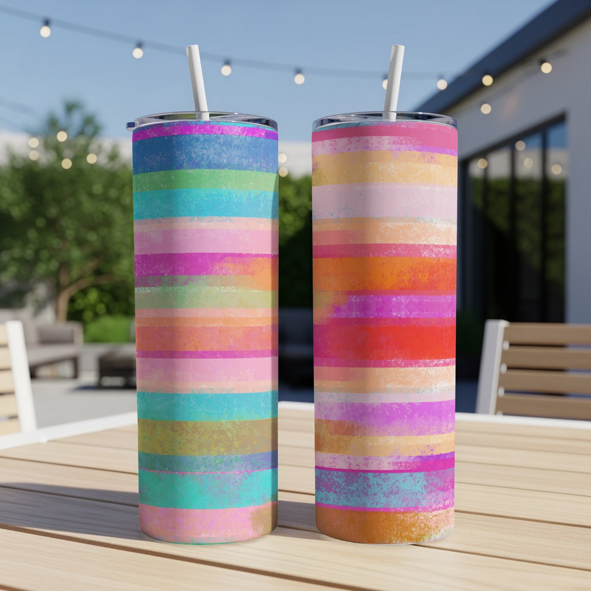 Two tumblers with colorful horizontal stripe patterns and white straws on a wooden table outdoors.